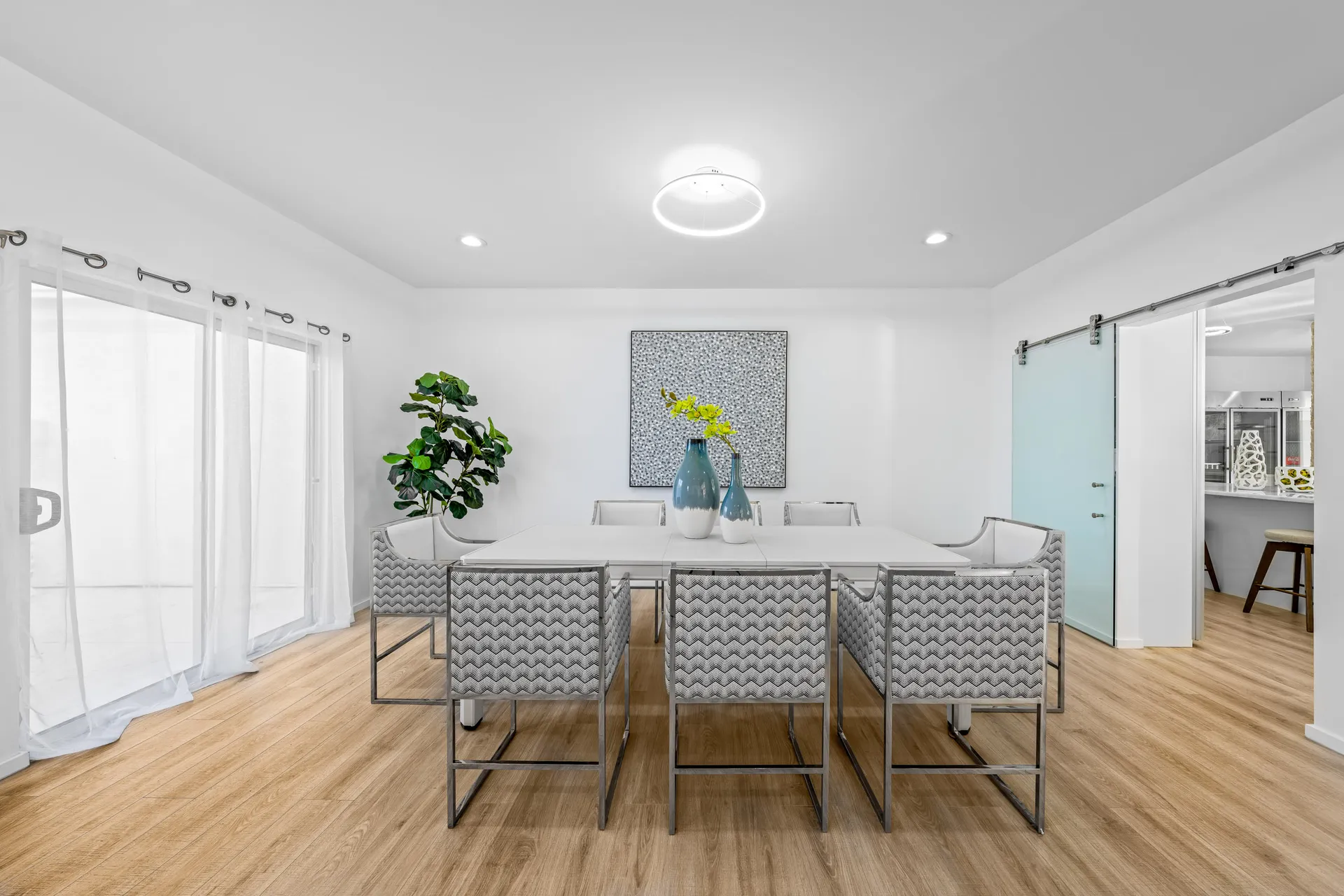 A white dining table with gray patterned chairs, teal vases at the center, and an abstract painting on the wall in a bright room.