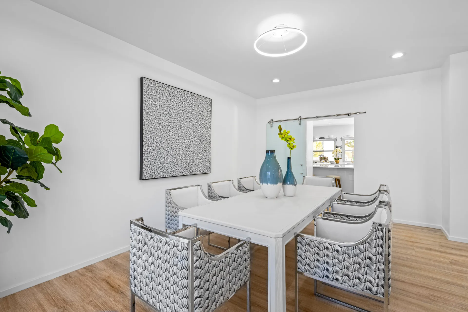 A bright dining room with a white table, silver patterned chairs, teal vases, and an abstract wall art piece.