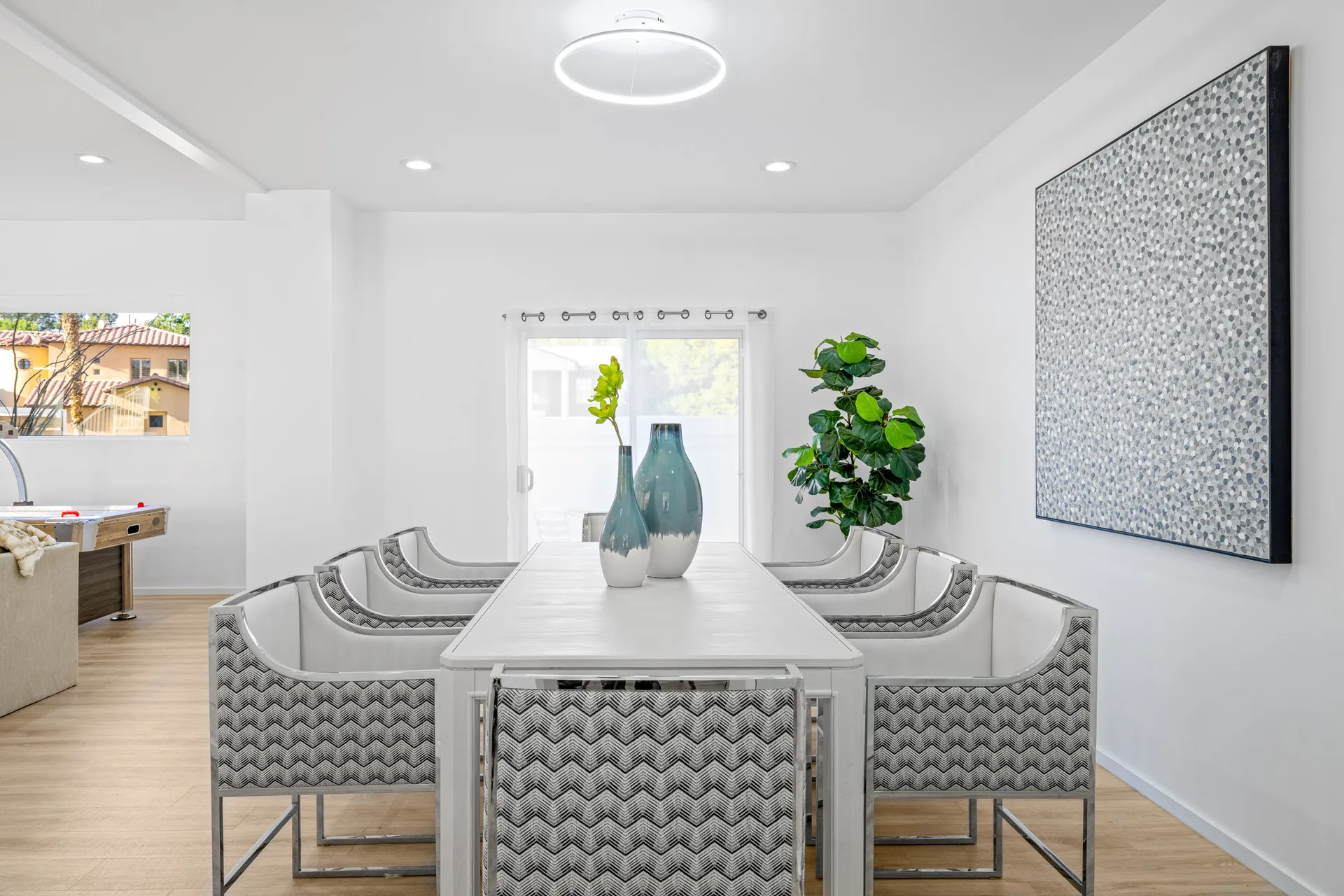 A white dining table with patterned chairs and teal vases, set in a bright room with large abstract art.