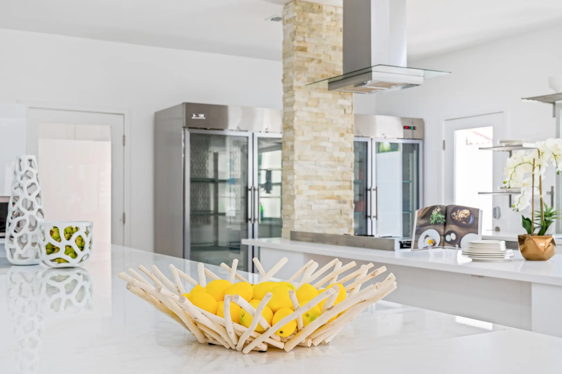A bright, modern kitchen with stainless steel appliances, a stone column, and a decorative white bowl filled with lemons on a sleek countertop.