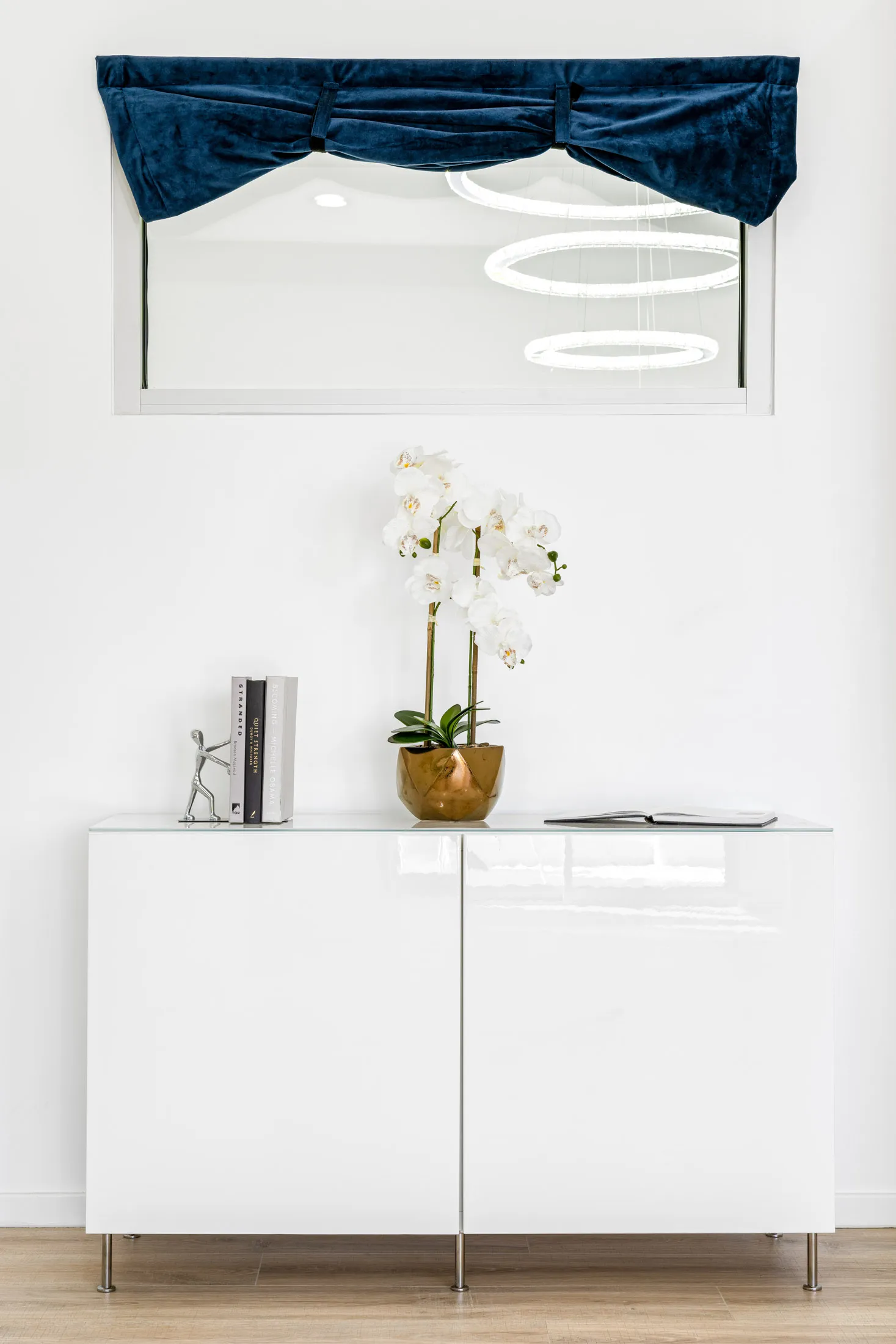 A glossy white console with a potted orchid in a gold vase, alongside books and a small silver figurine.