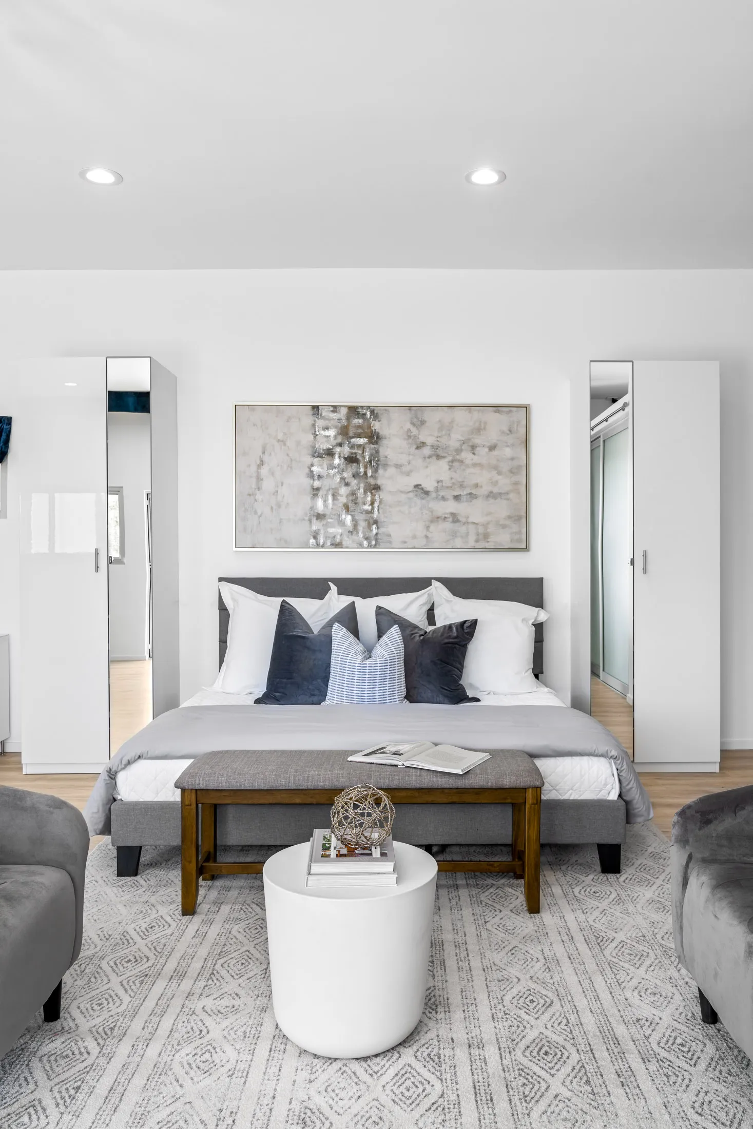 A contemporary bedroom featuring a gray bed with layered pillows, a wooden bench at the foot, and an abstract painting overhead.
