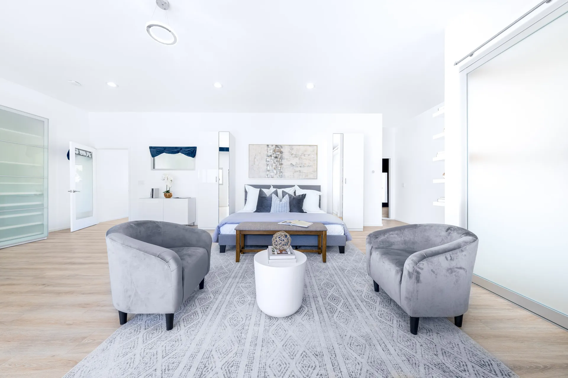 A bright, white bedroom with a gray bed, two gray chairs, and a wooden bench on a patterned rug.