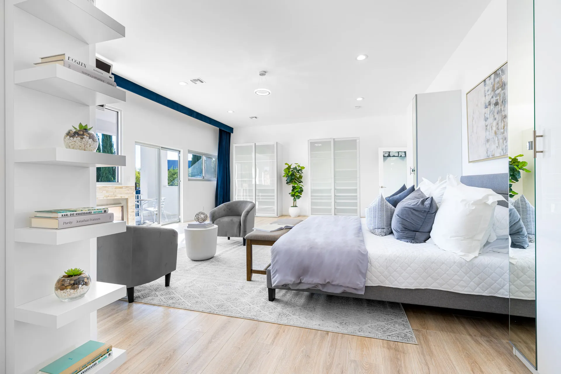 A bright, open bedroom with a gray bed, floating shelves, and a cozy seating area near large windows.