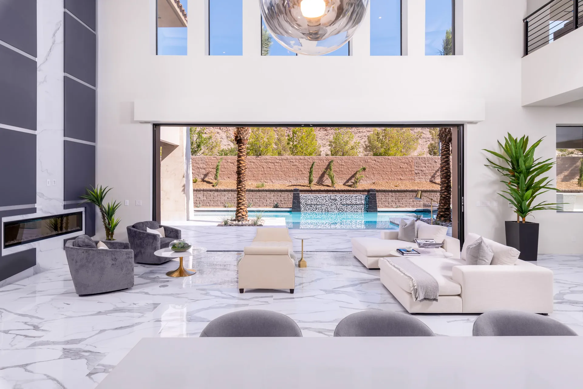 An indoor living space with a white marble floor, high ceilings, and large windows overlooking a pool and palm trees. The space is furnished with white sofas, a glass coffee table, and a dining area. The design is minimalist and luxurious, creating a serene and inviting atmosphere.