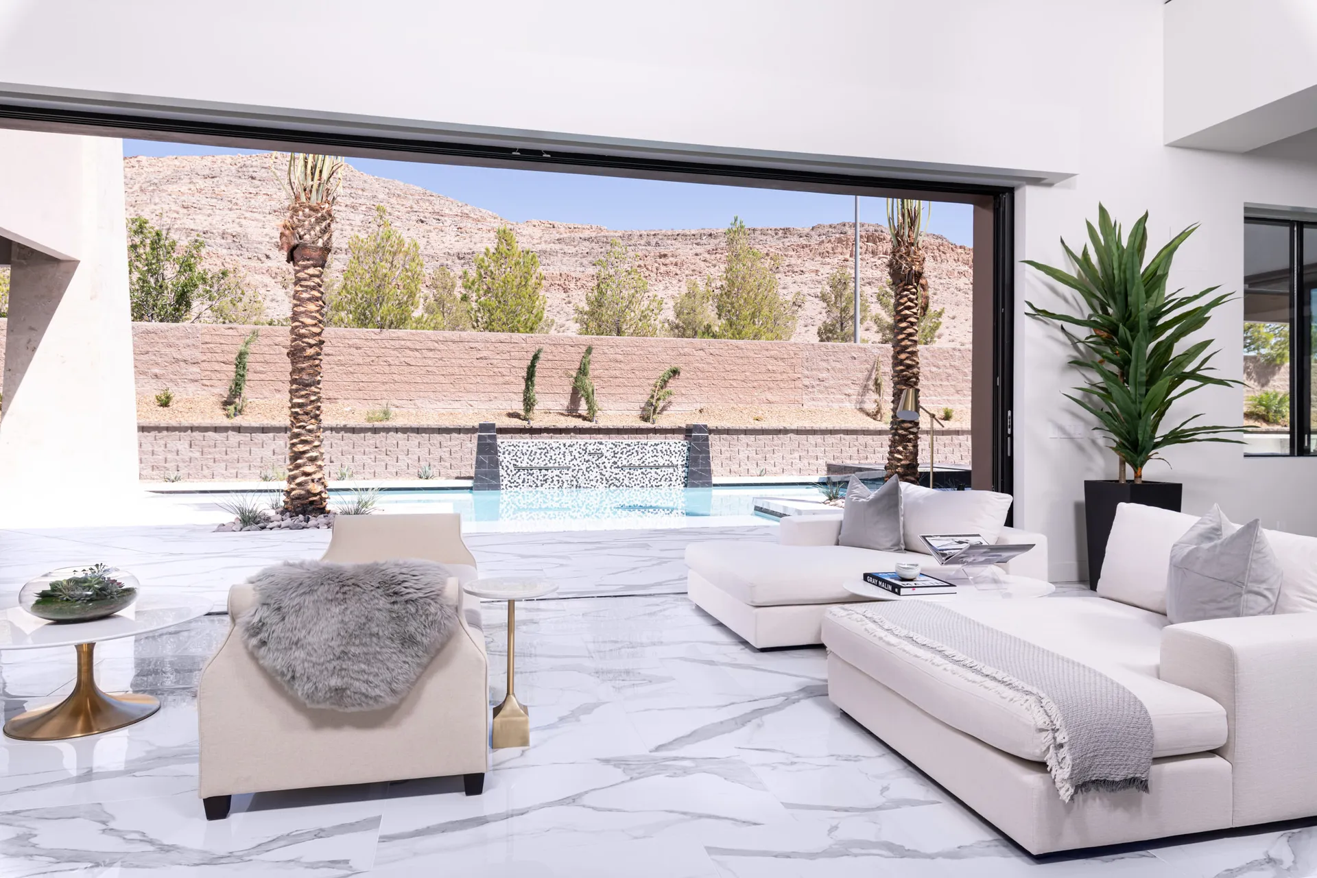 An outdoor living space with a white marble floor, white sofas, and a view of a pool and palm trees. The space features a minimalist design and a luxurious atmosphere, creating a serene and inviting outdoor experience.