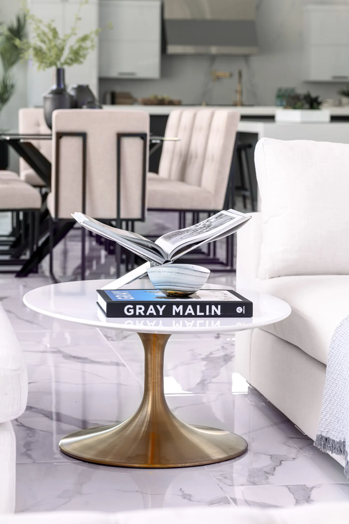 A close-up of a white coffee table with a brass base, adorned with a book titled "GRAY MALIN" and an open magazine. The background features a modern living room with a neutral color palette, creating a sophisticated and inviting atmosphere.