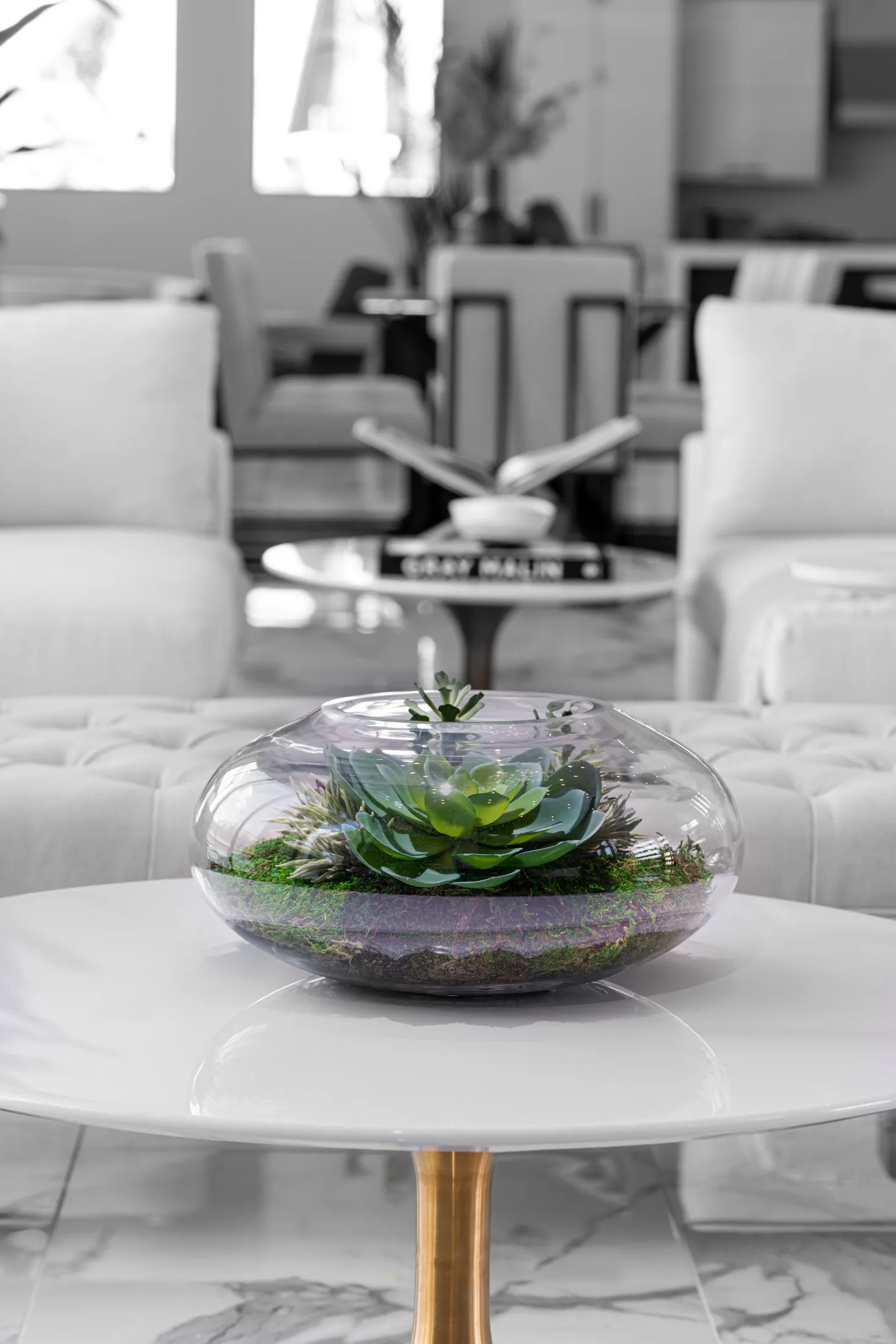 A close-up of a glass terrarium with succulents, placed on a white coffee table. The background features a modern living room with a neutral color palette, creating a serene and inviting atmosphere.
