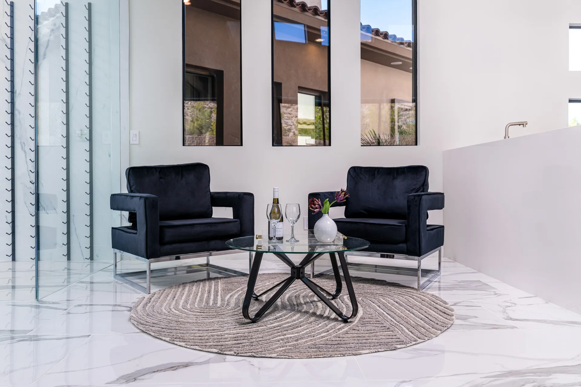 A modern living room with a glass coffee table, two black armchairs, and a round rug. The table is adorned with a wine bottle, glasses, and a vase with flowers, creating a sophisticated and inviting atmosphere.