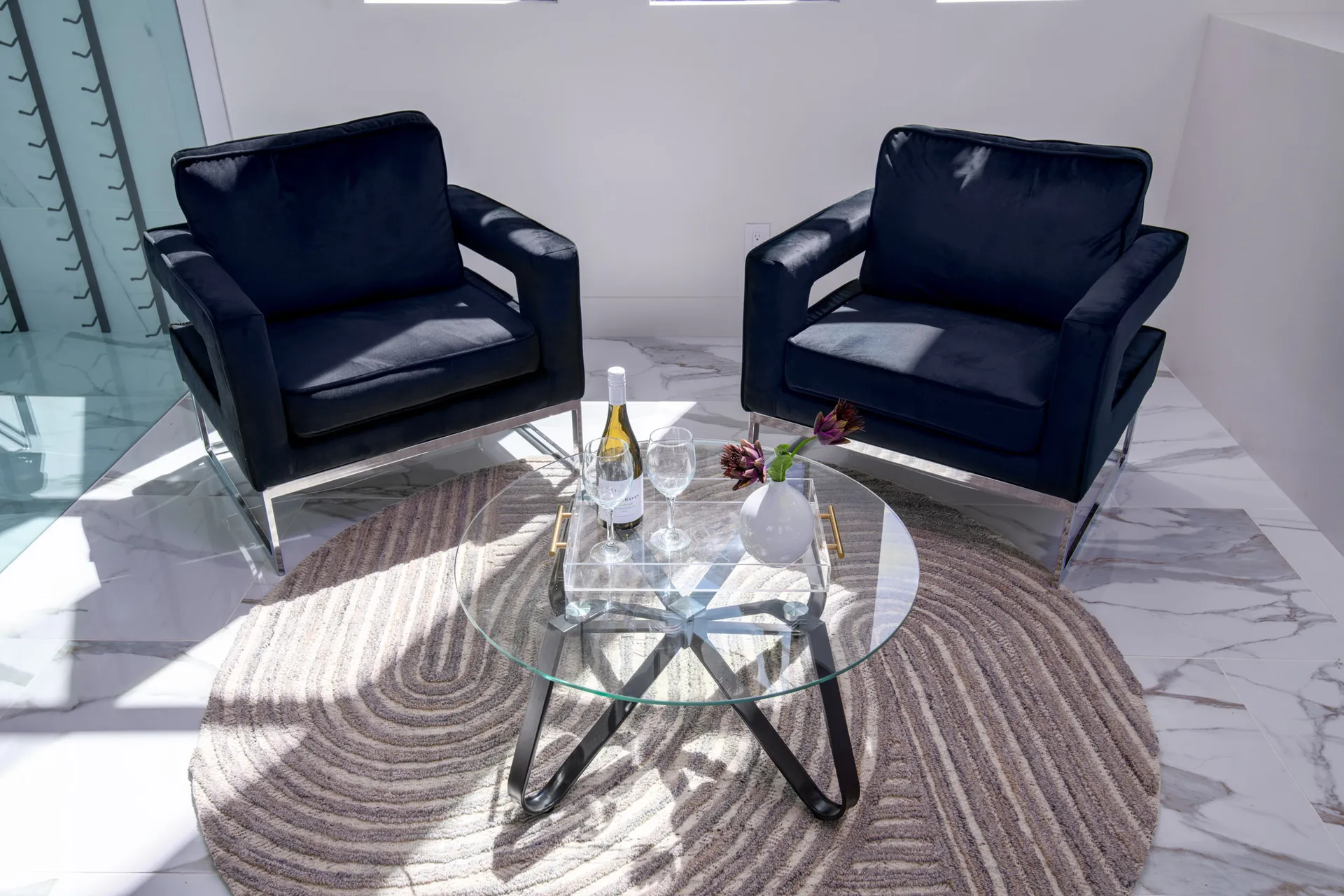 A modern living room with a glass coffee table, two black armchairs, and a round rug. The table is adorned with a wine bottle, glasses, and a vase with flowers, creating a sophisticated and inviting atmosphere.