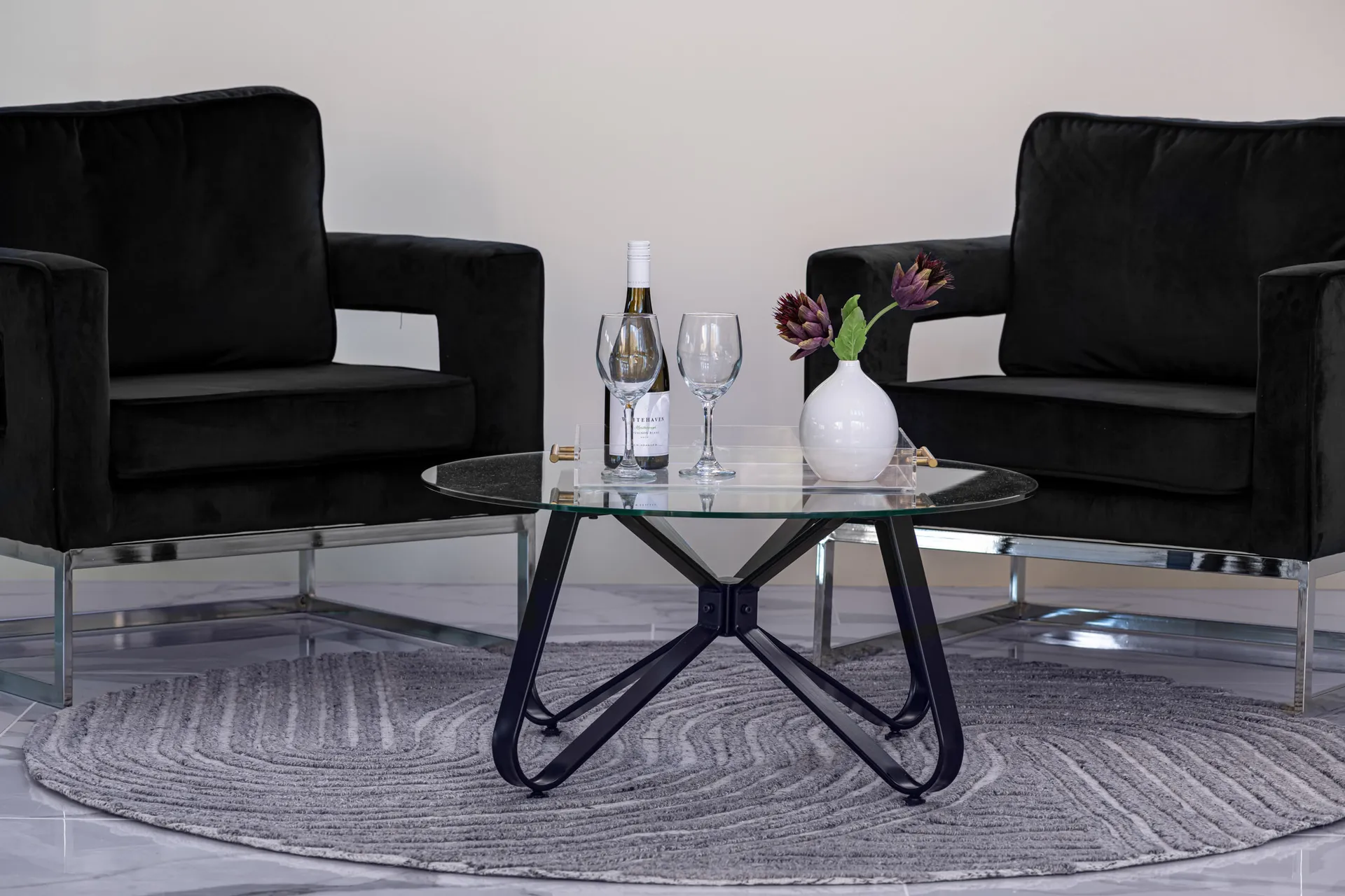 A modern living room with a glass coffee table, two black armchairs, and a round rug. The table is adorned with a wine bottle, glasses, and a vase with flowers, creating a sophisticated and inviting atmosphere.
