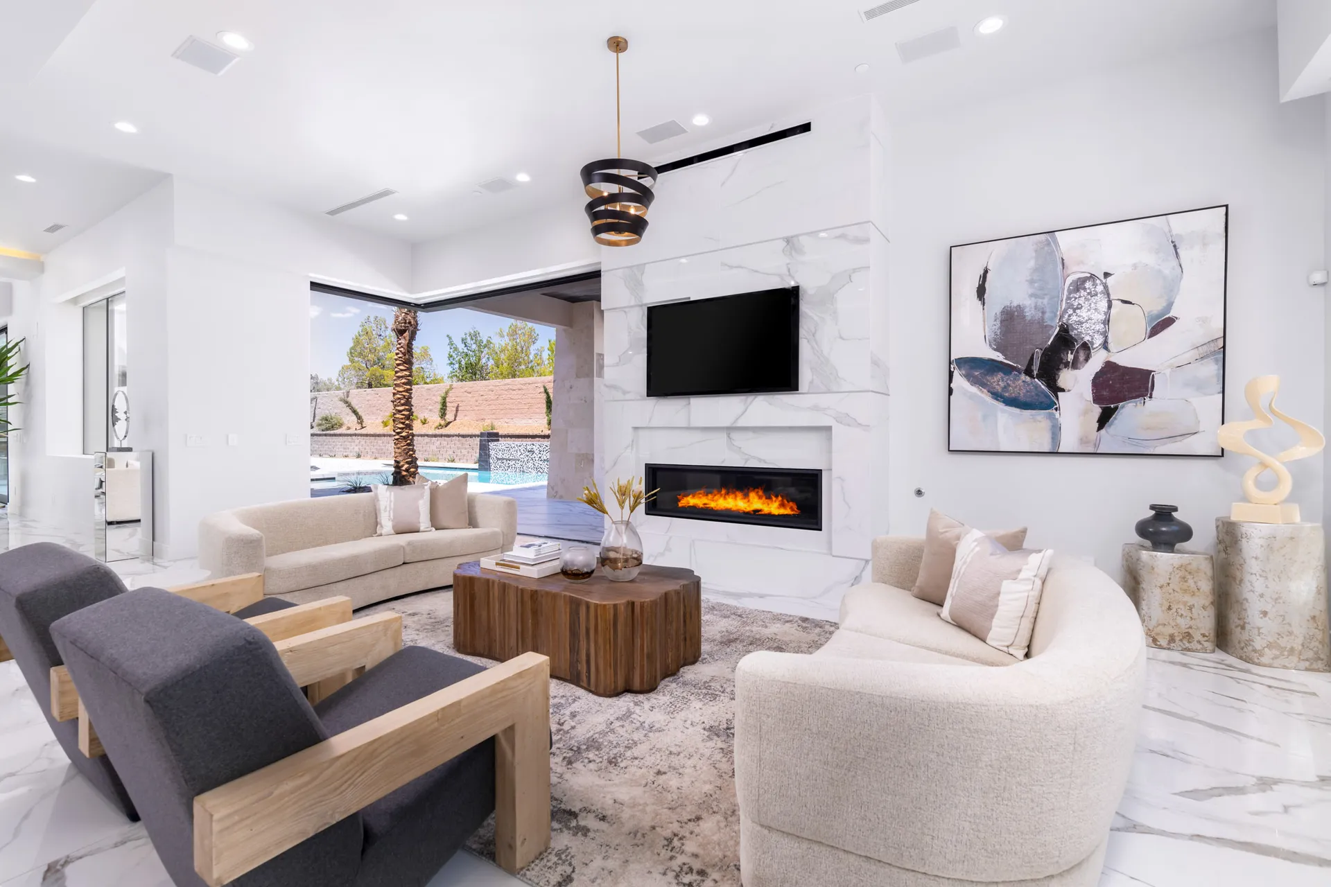 High-End Home Staging A modern living room with a fireplace, a large window, and a view of an outdoor pool area. The room features a neutral color palette, with a mix of gray and beige furniture, and a wooden coffee table. The design is contemporary, with clean lines and a minimalist aesthetic.