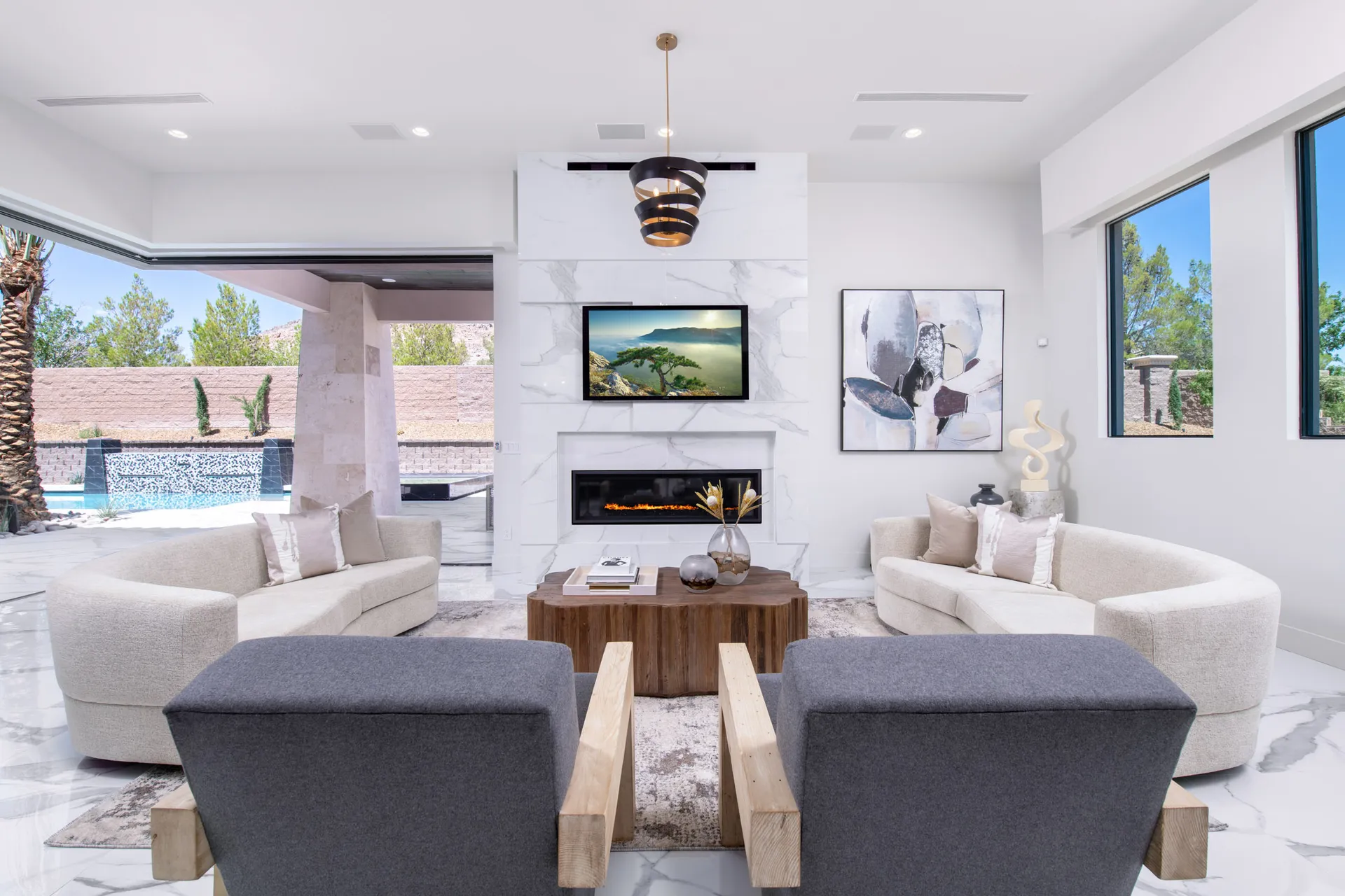 A modern living room with a fireplace, a large window, and a view of an outdoor pool area. The room features a neutral color palette, with a mix of gray and beige furniture, and a wooden coffee table. The design is contemporary, with clean lines and a minimalist aesthetic.