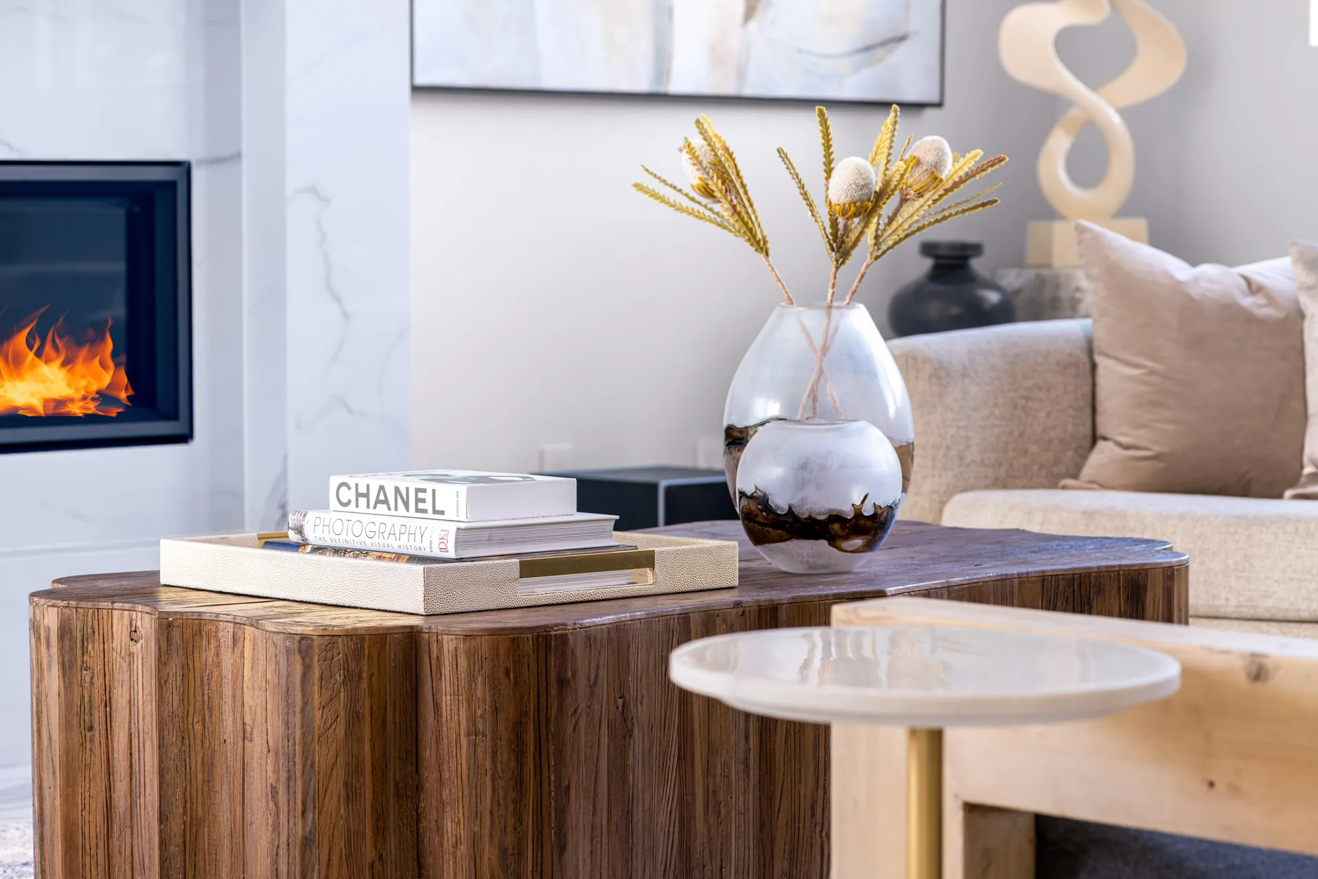 A close-up of a wooden coffee table with decorative vases, books, and a plant in the background. The design features a modern aesthetic with clean lines and a neutral color palette, creating a sophisticated and elegant look.