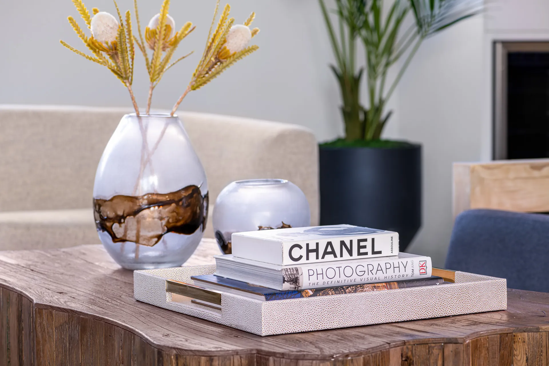 A close-up of a wooden coffee table with decorative vases, books, and a plant in the background. The design features a modern aesthetic with clean lines and a neutral color palette, creating a sophisticated and elegant look.