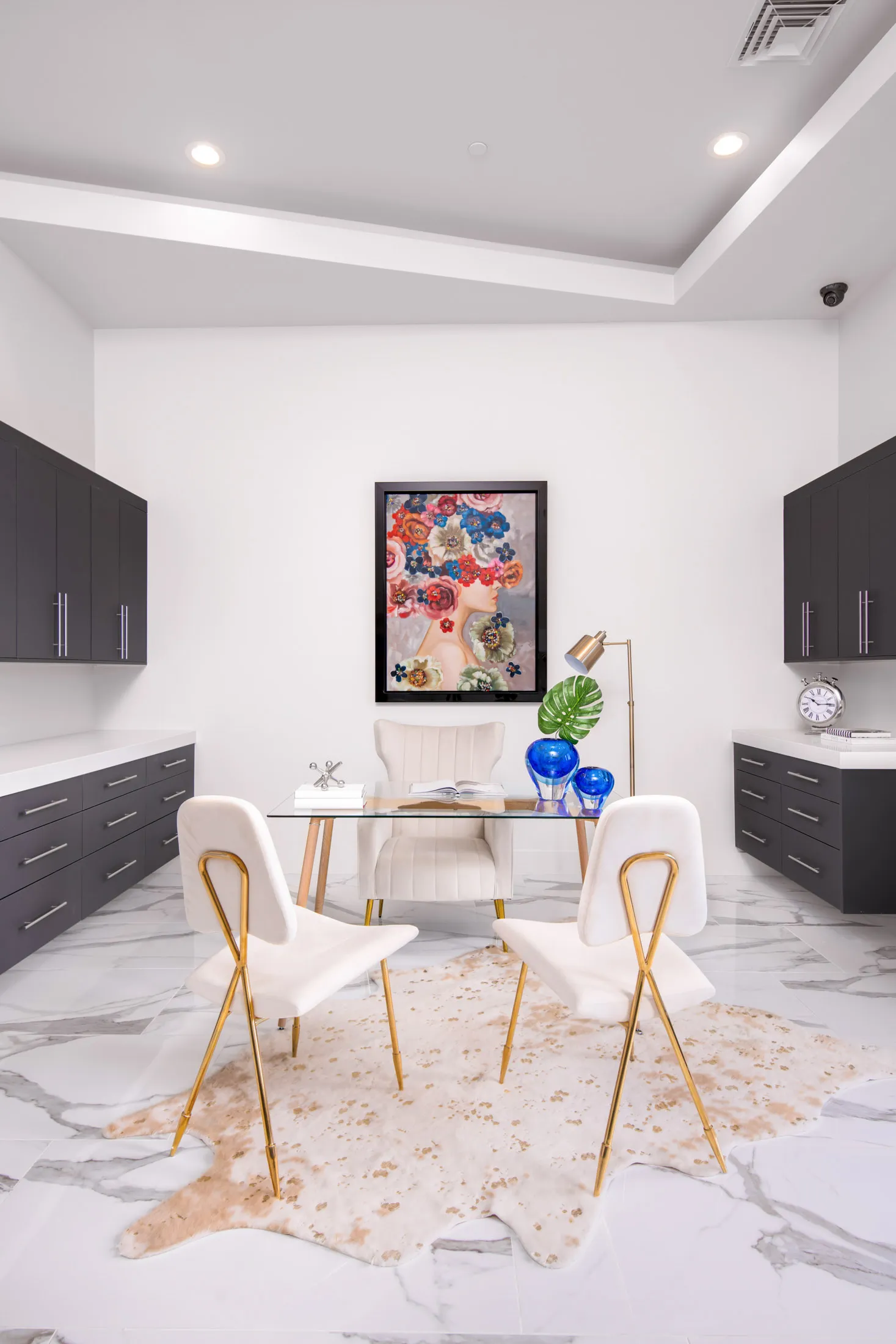 A home office with a glass desk, white chairs, a colorful artwork on the wall, and decorative objects on the desk. The design features a modern aesthetic with clean lines and a neutral color palette, creating a sophisticated and elegant look.