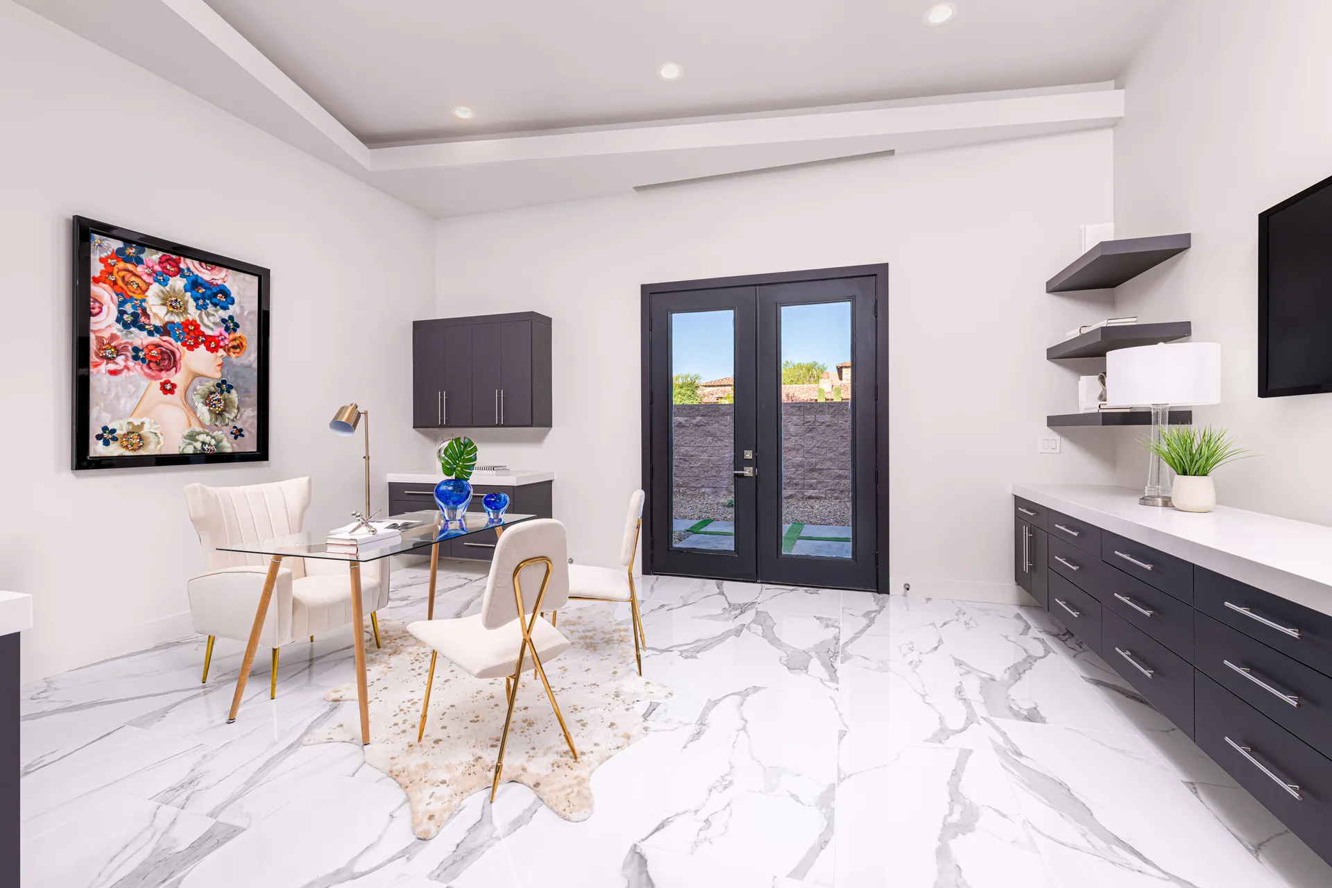 A home office with a glass desk, white chairs, a colorful artwork on the wall, and decorative objects on the desk. The design features a modern aesthetic with clean lines and a neutral color palette, creating a sophisticated and elegant look.
