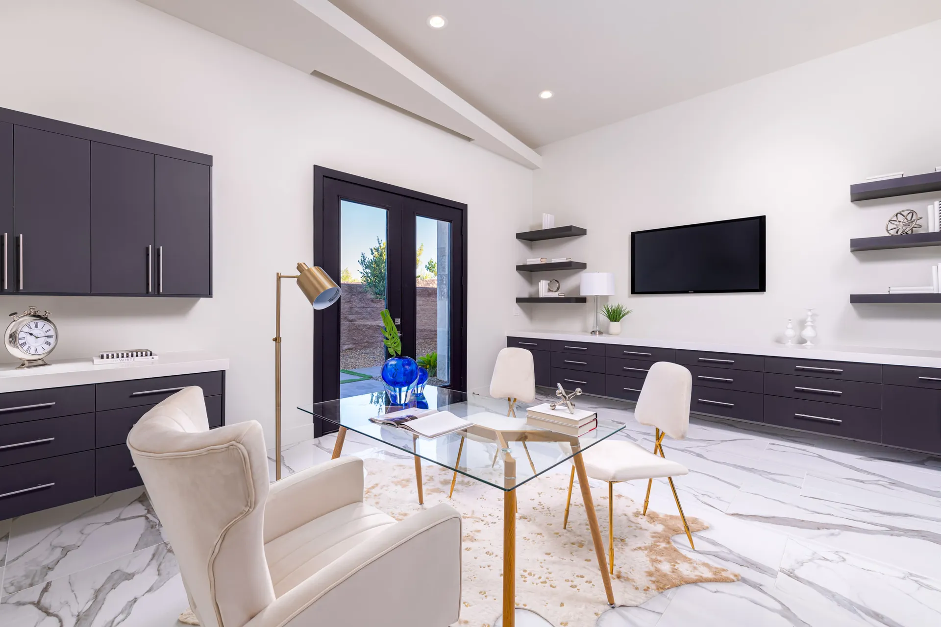 A home office with a glass desk, white chairs, a colorful artwork on the wall, and decorative objects on the desk. The design features a modern aesthetic with clean lines and a neutral color palette, creating a sophisticated and elegant look.