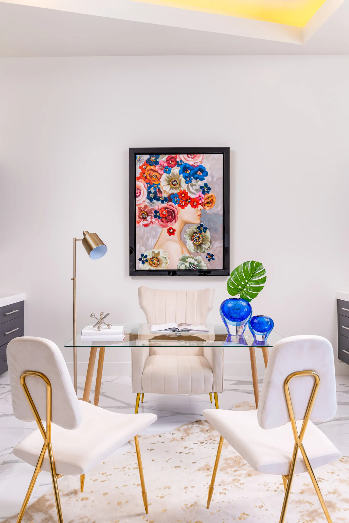 A home office with a glass desk, white chairs, a colorful artwork on the wall, and decorative objects on the desk. The design features a modern aesthetic with clean lines and a neutral color palette, creating a sophisticated and elegant look.