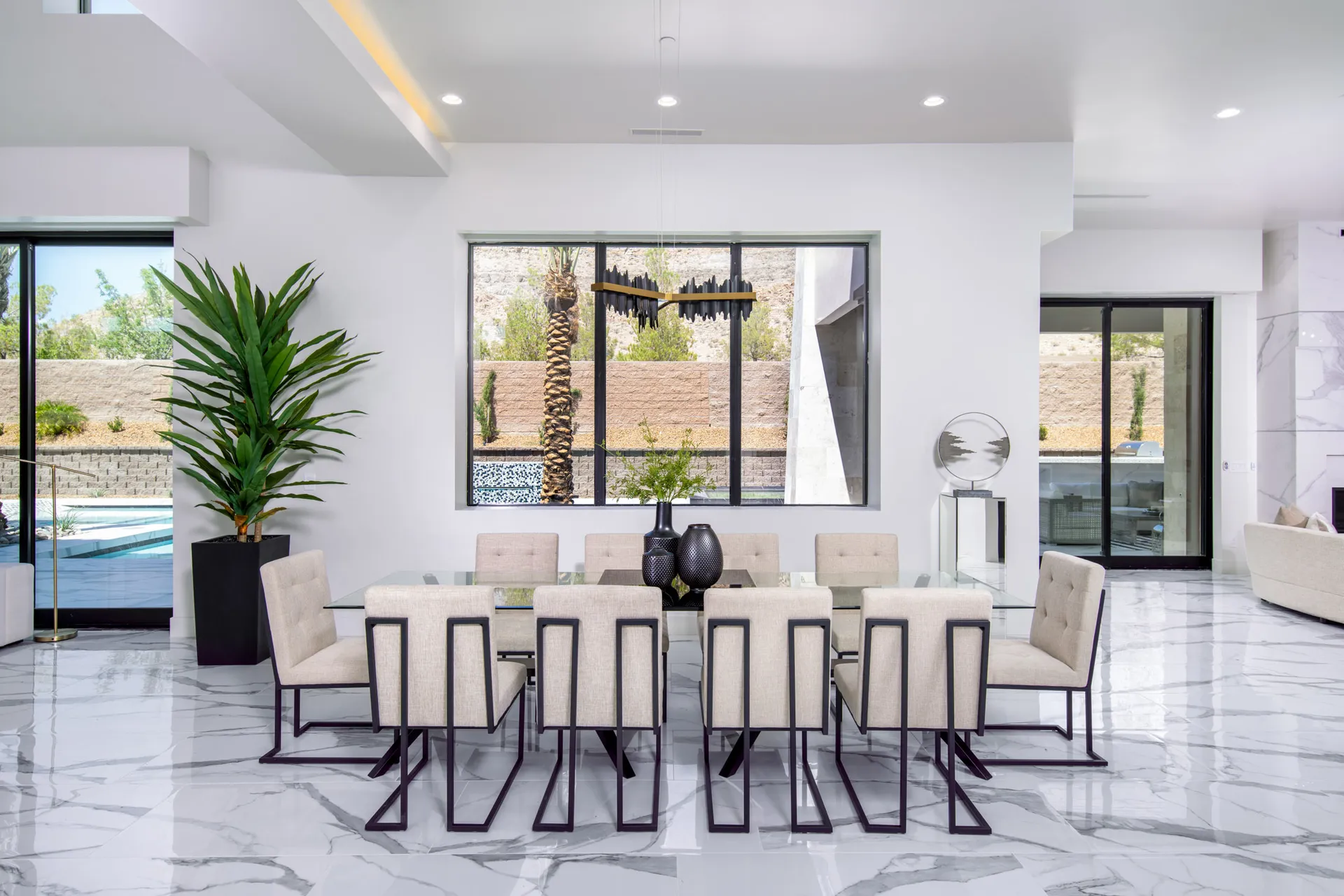 A dining room with beige upholstered chairs, a glass table, decorative vases with greenery, and large windows offering a view of the outdoors. The design features a modern aesthetic with clean lines and a neutral color palette, creating a sophisticated and elegant look.