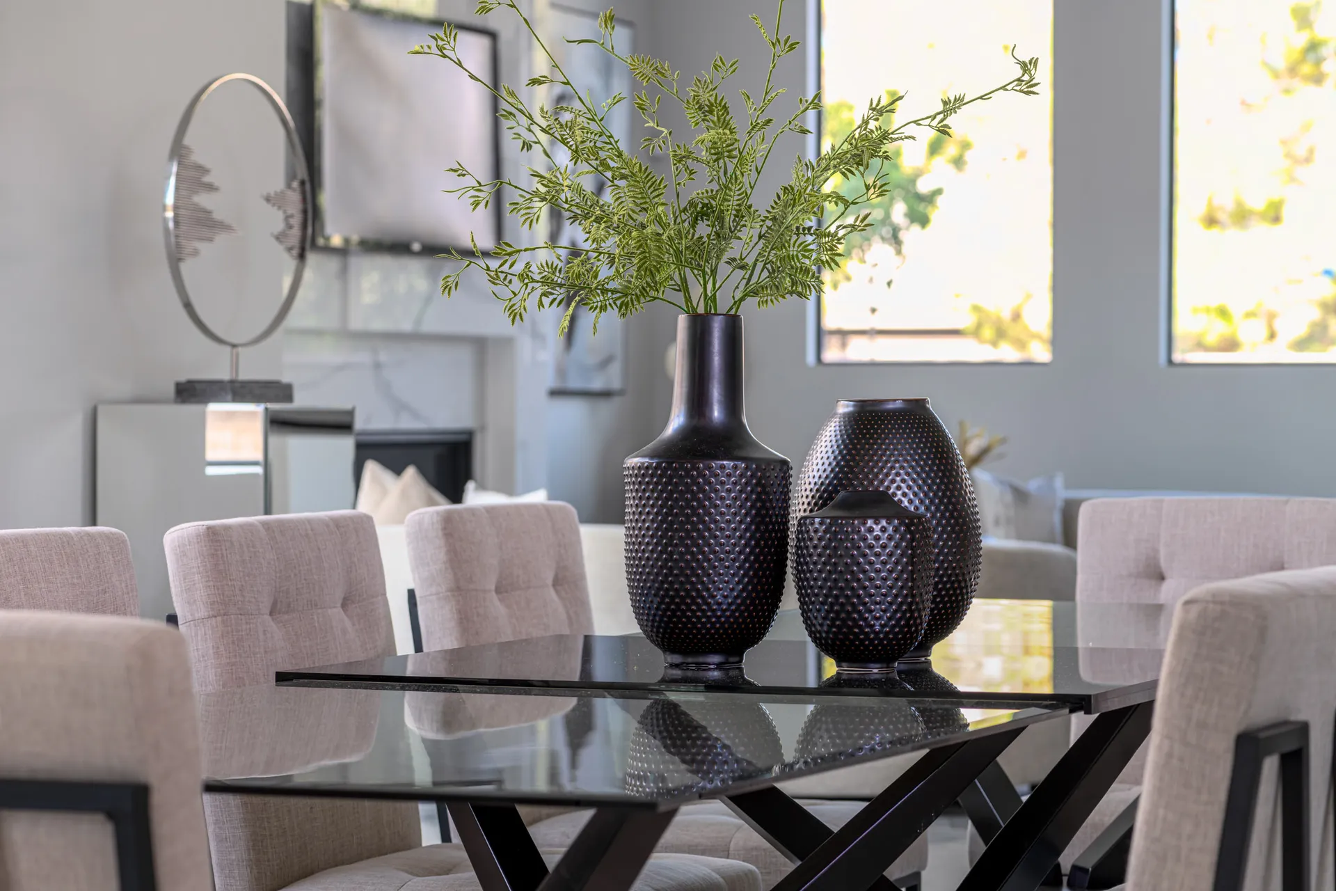 A dining room with a glass table, beige upholstered chairs, and decorative vases with greenery. The design features a modern aesthetic with clean lines and a neutral color palette, creating a sophisticated and elegant look.