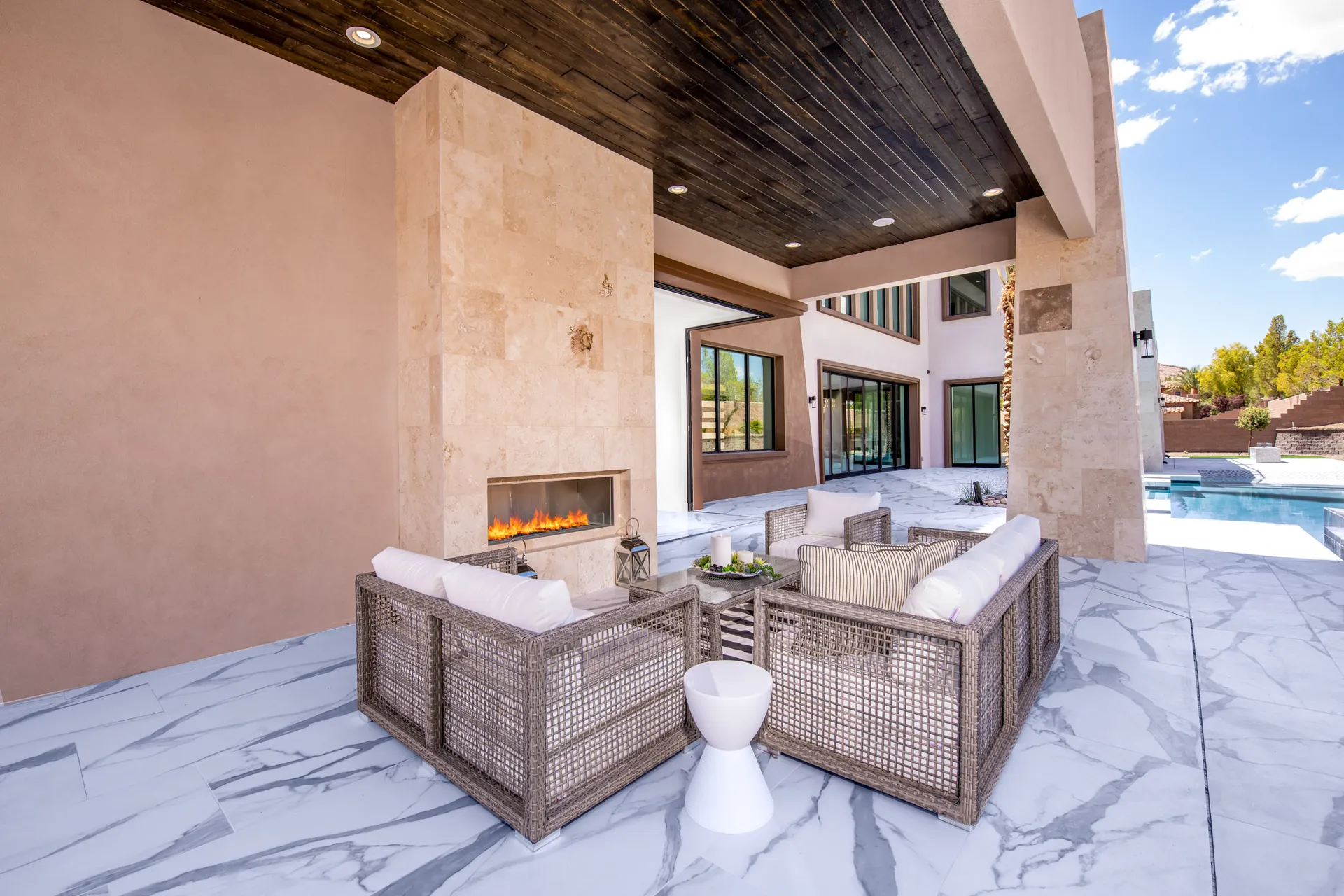 An outdoor seating area with wicker furniture, cushions, a stone fireplace, and a view of a pool and palm trees in the background. The arrangement features a comfortable and inviting setting, perfect for relaxation and outdoor gatherings.