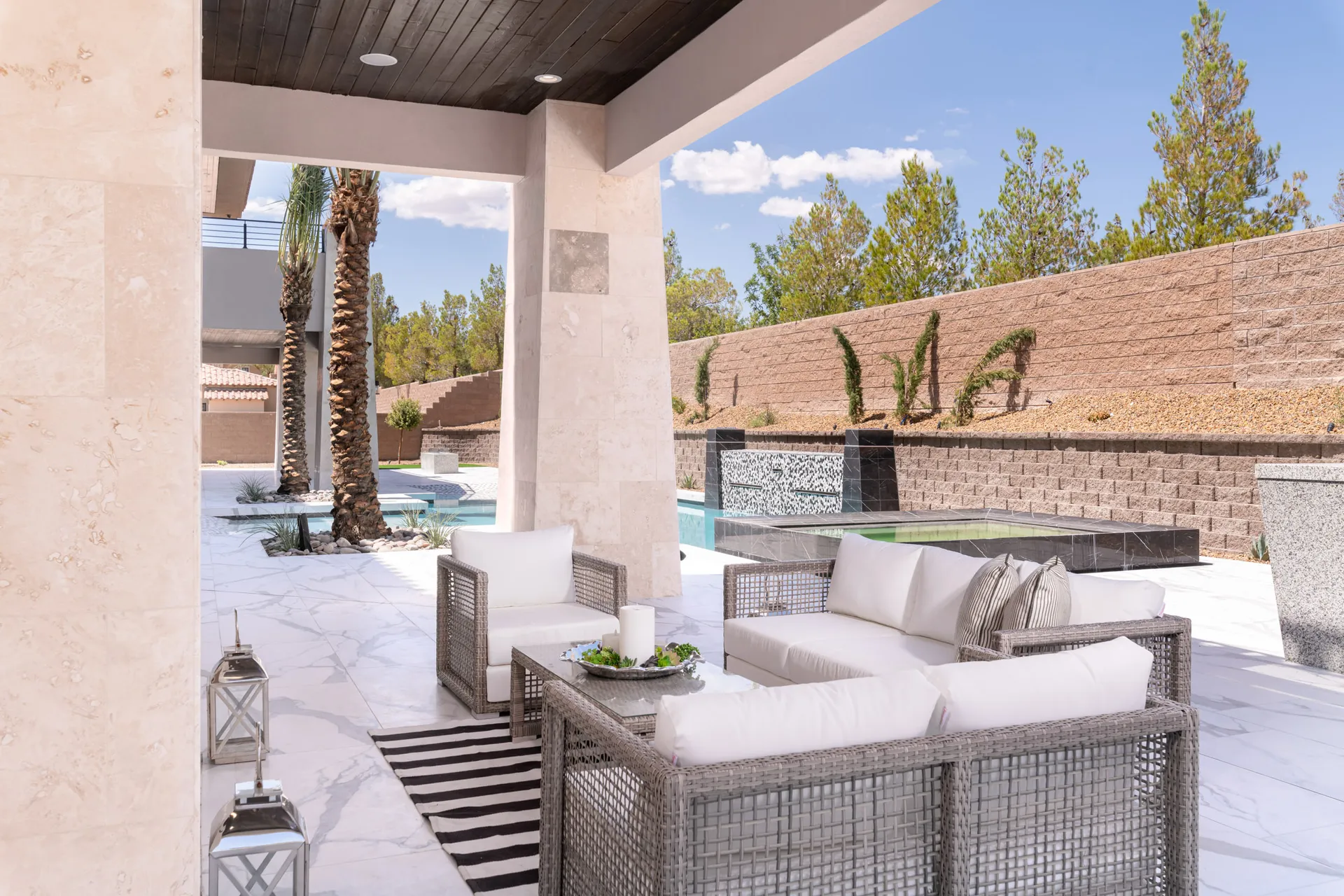 An outdoor seating area with wicker furniture, cushions, and a view of a pool and palm trees in the background. The arrangement features a comfortable and inviting setting, perfect for relaxation and outdoor gatherings.