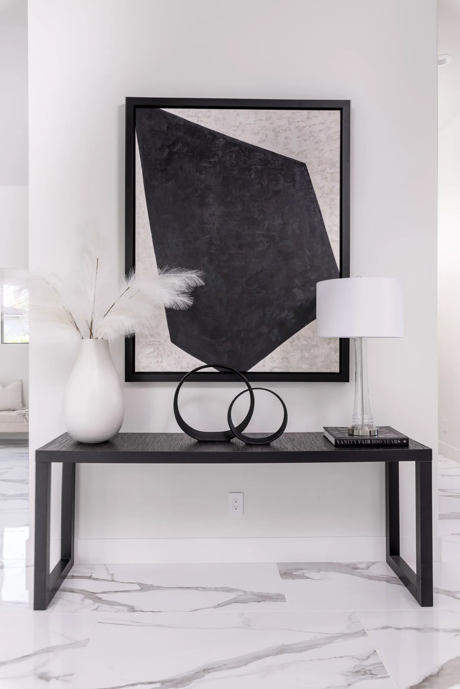 A decorative console table with a large abstract painting, a white vase with pampas grass, and a table lamp. The design features a modern aesthetic with clean lines and a neutral color palette, creating a sophisticated and elegant look.