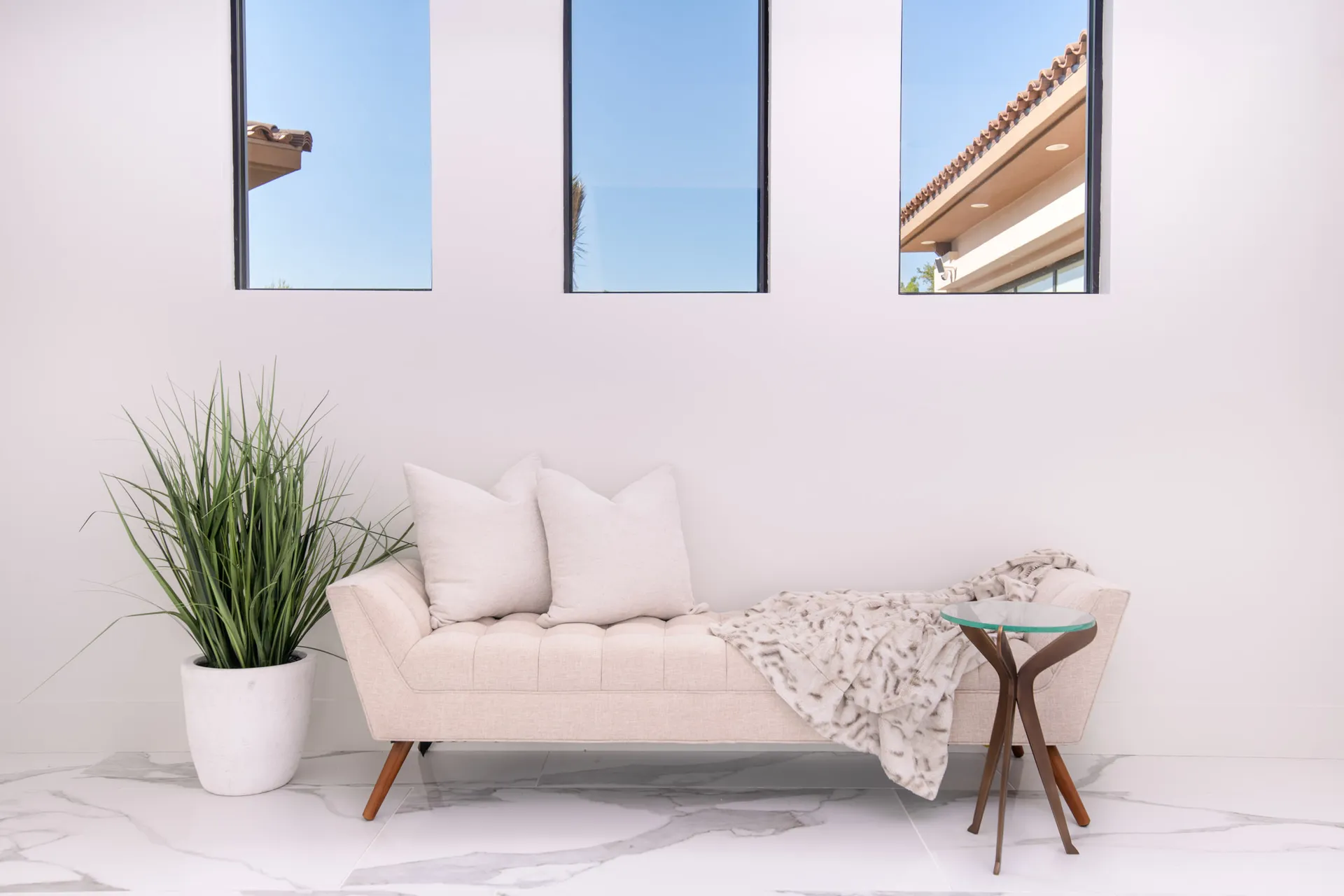 A hallway with a beige bench, a throw blanket, and a potted plant, with large windows in the background. The design features a minimalist aesthetic with clean lines and a neutral color palette, creating a serene and inviting atmosphere.