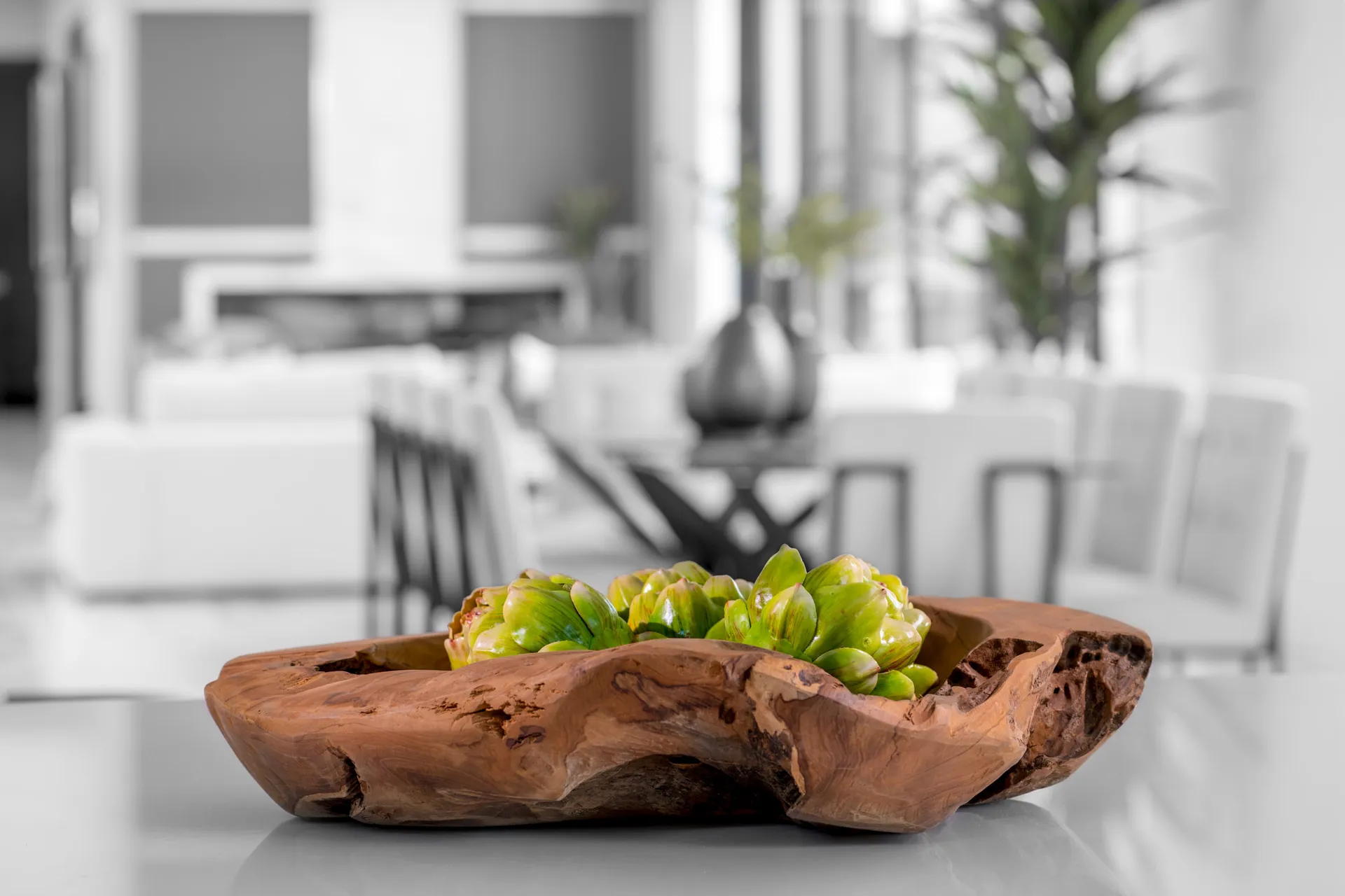 A wooden bowl with succulents, placed on a reflective surface with a blurred background of a modern interior. The arrangement features a mix of textures and colors, creating a visually appealing and natural design.