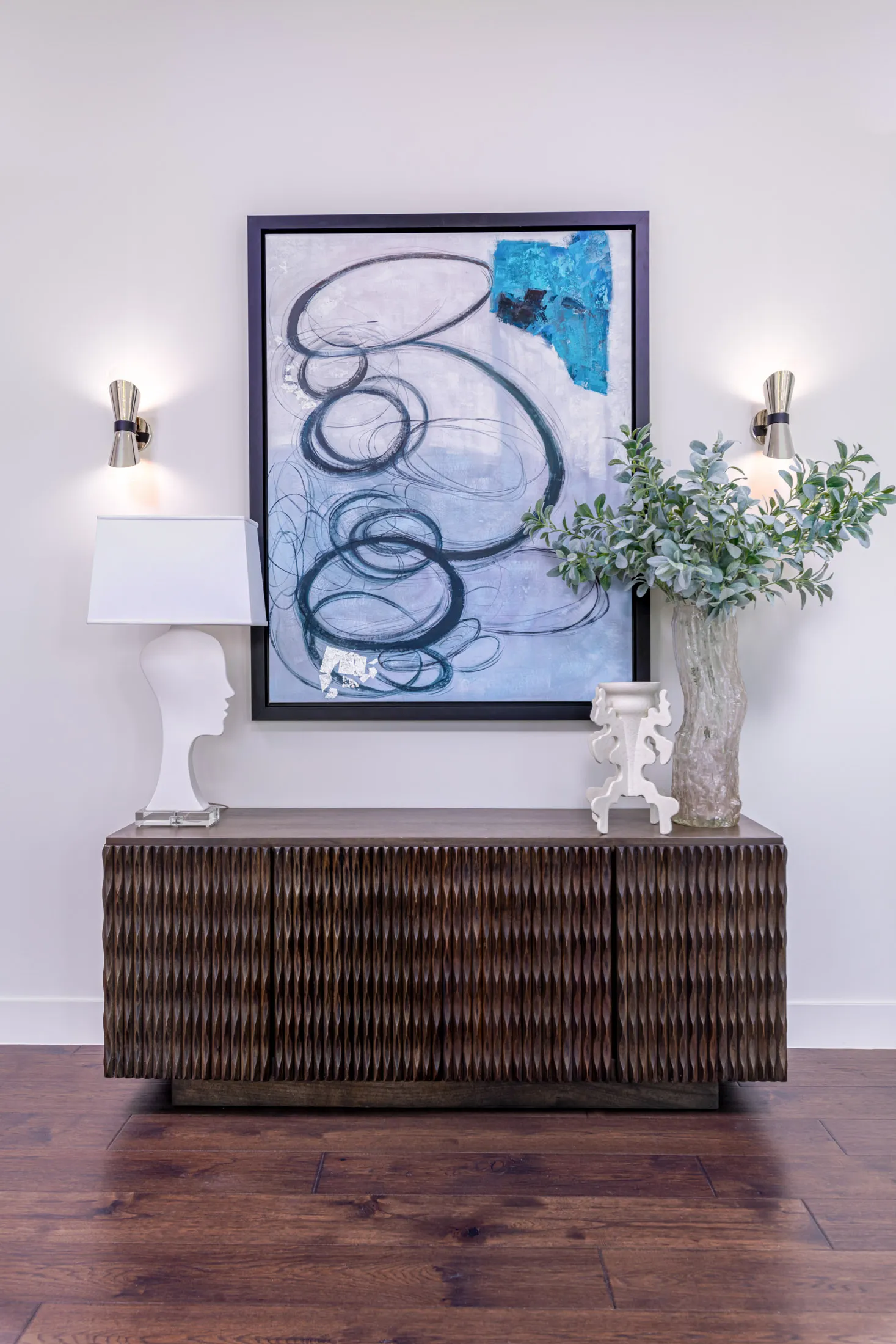 A decorative console table with a large abstract painting, a table lamp, and a vase with green foliage. The design features a modern aesthetic with clean lines and a neutral color palette, creating a sophisticated and elegant look.