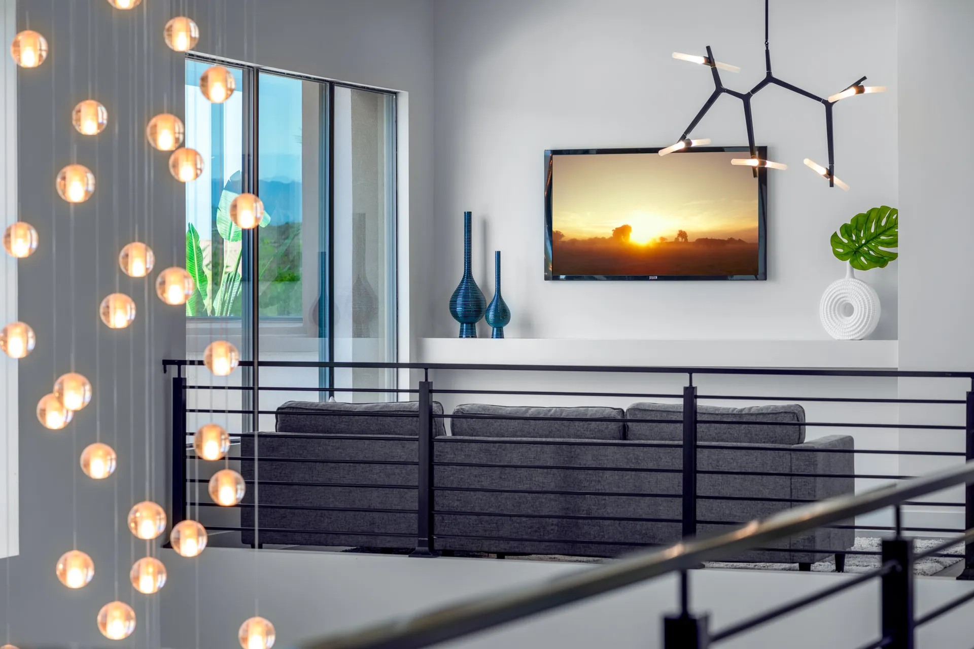 A loft area with a modern chandelier, a sofa, and large windows offering a view of the outdoors. The design features a modern aesthetic with clean lines and a neutral color palette, creating a sophisticated and inviting atmosphere.