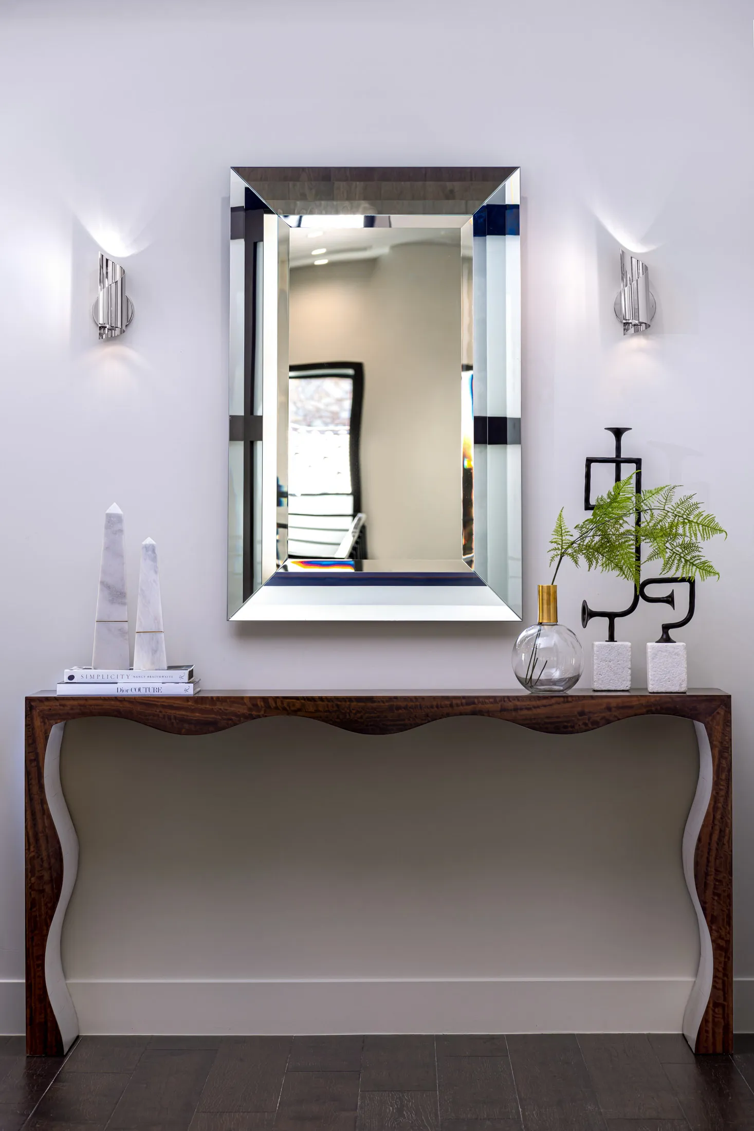 A decorative console table with a mirror, marble obelisks, and a vase with green foliage. The design features a modern aesthetic with clean lines and a neutral color palette, creating a sophisticated and elegant look.