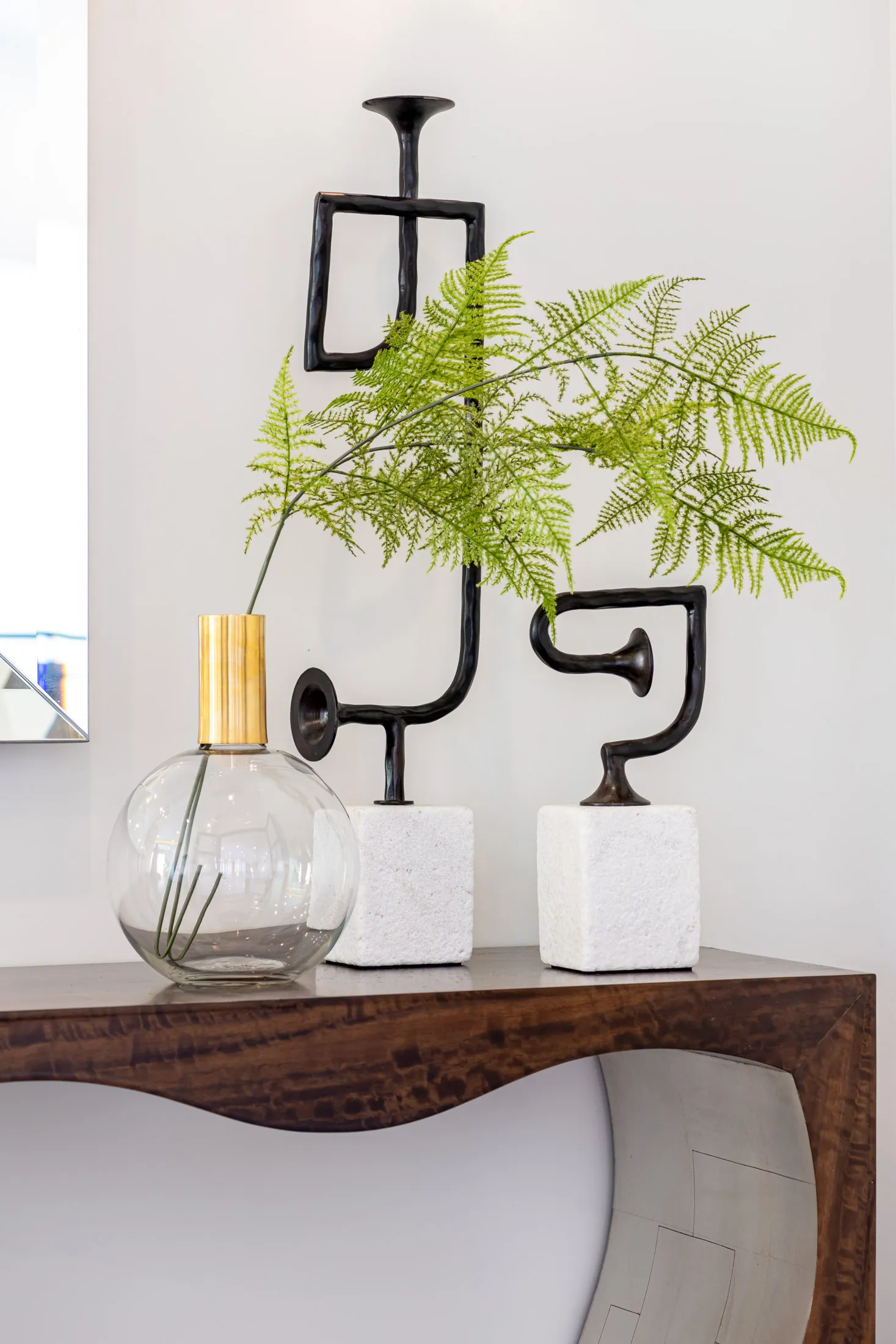 A decorative arrangement of a glass vase with green foliage and a modern sculpture on a wooden table. The design features a minimalist aesthetic with clean lines and a neutral color palette, creating a sophisticated and elegant look.