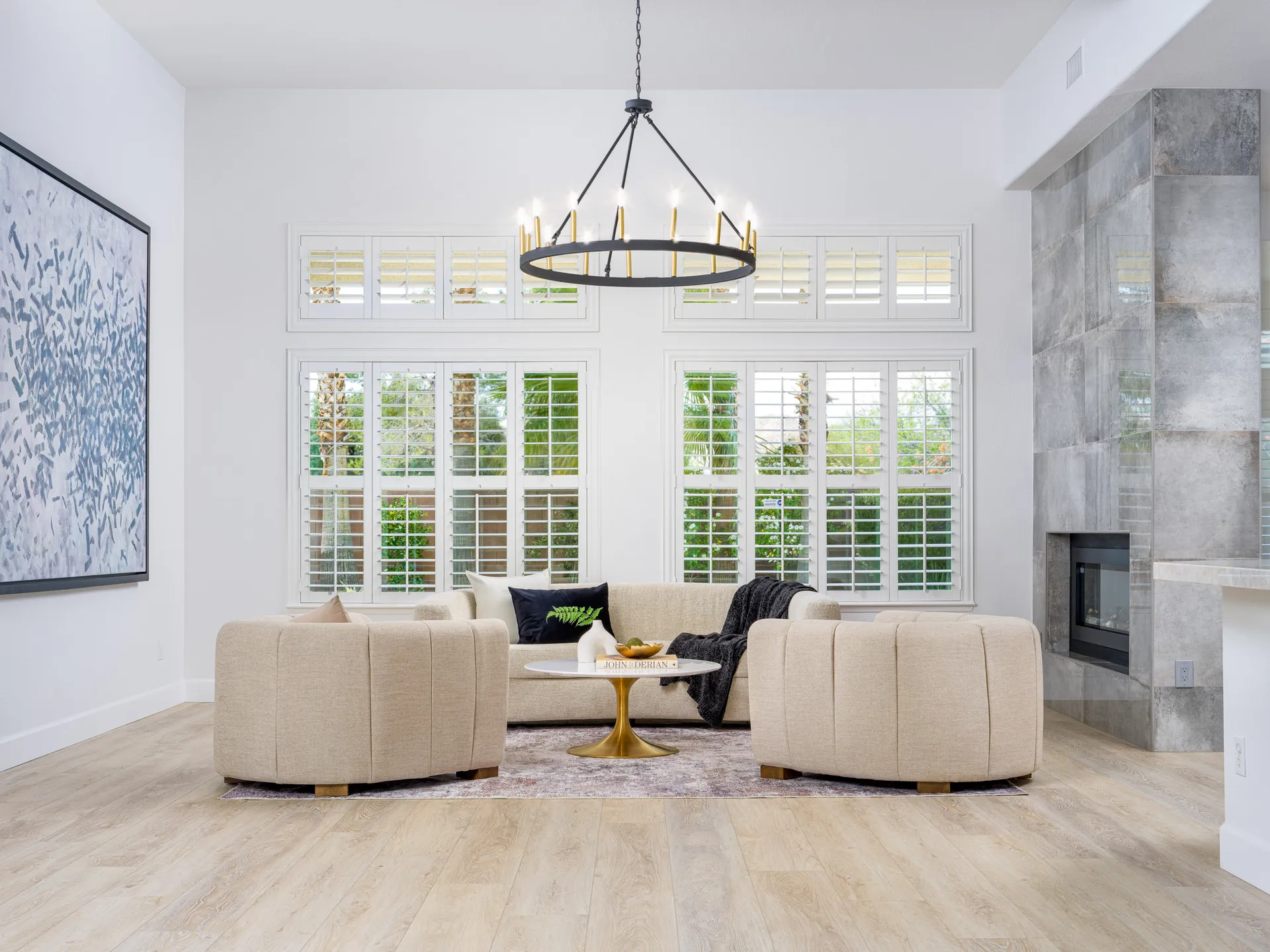 A living room with neutral curved chairs, a matching sofa, tall white shutters, and a statement chandelier near a tiled fireplace