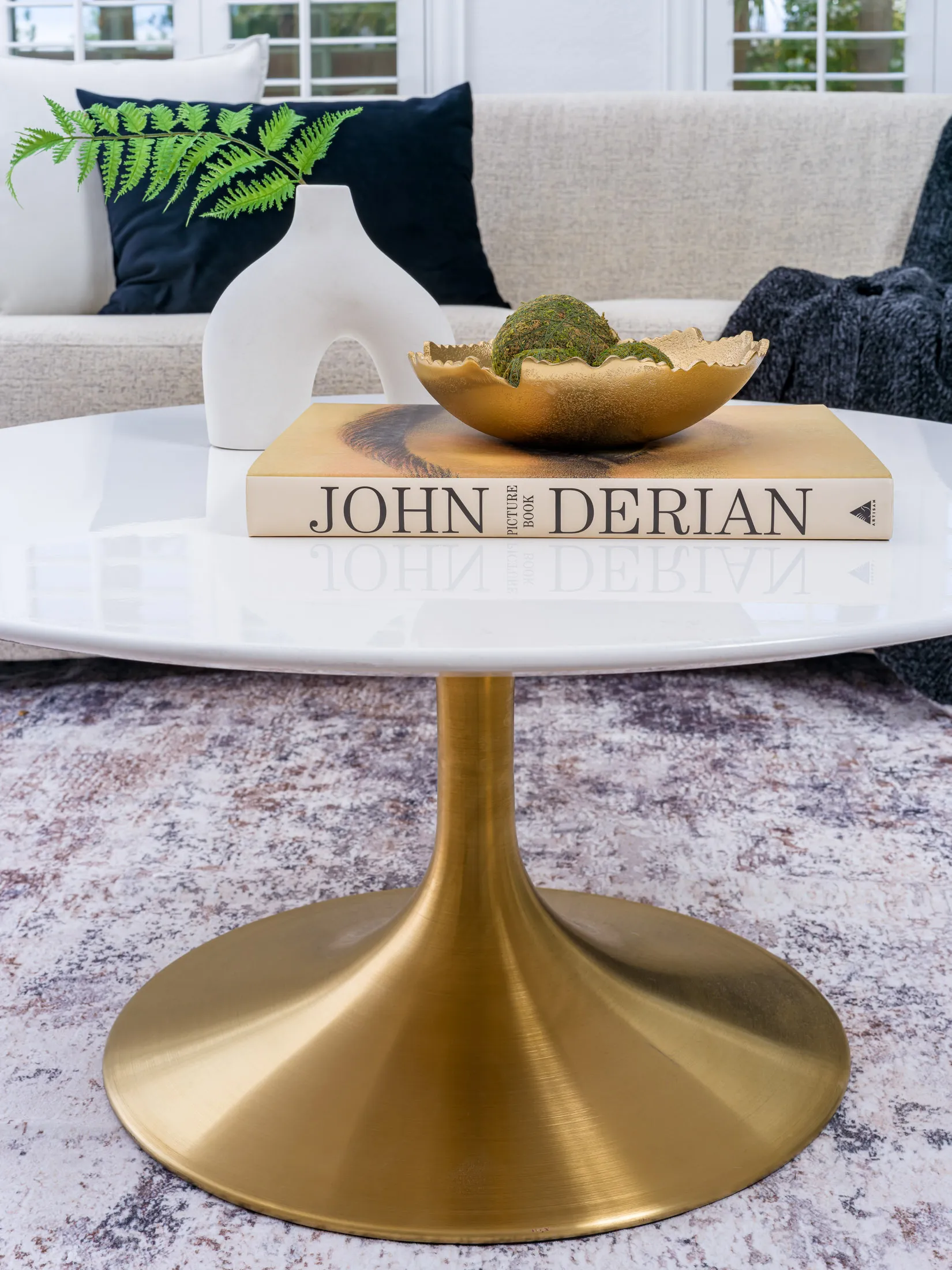 A white coffee table with a gold pedestal base, topped by a gold bowl of moss spheres on a hardcover book and a sculptural vase holding a fern leaf.