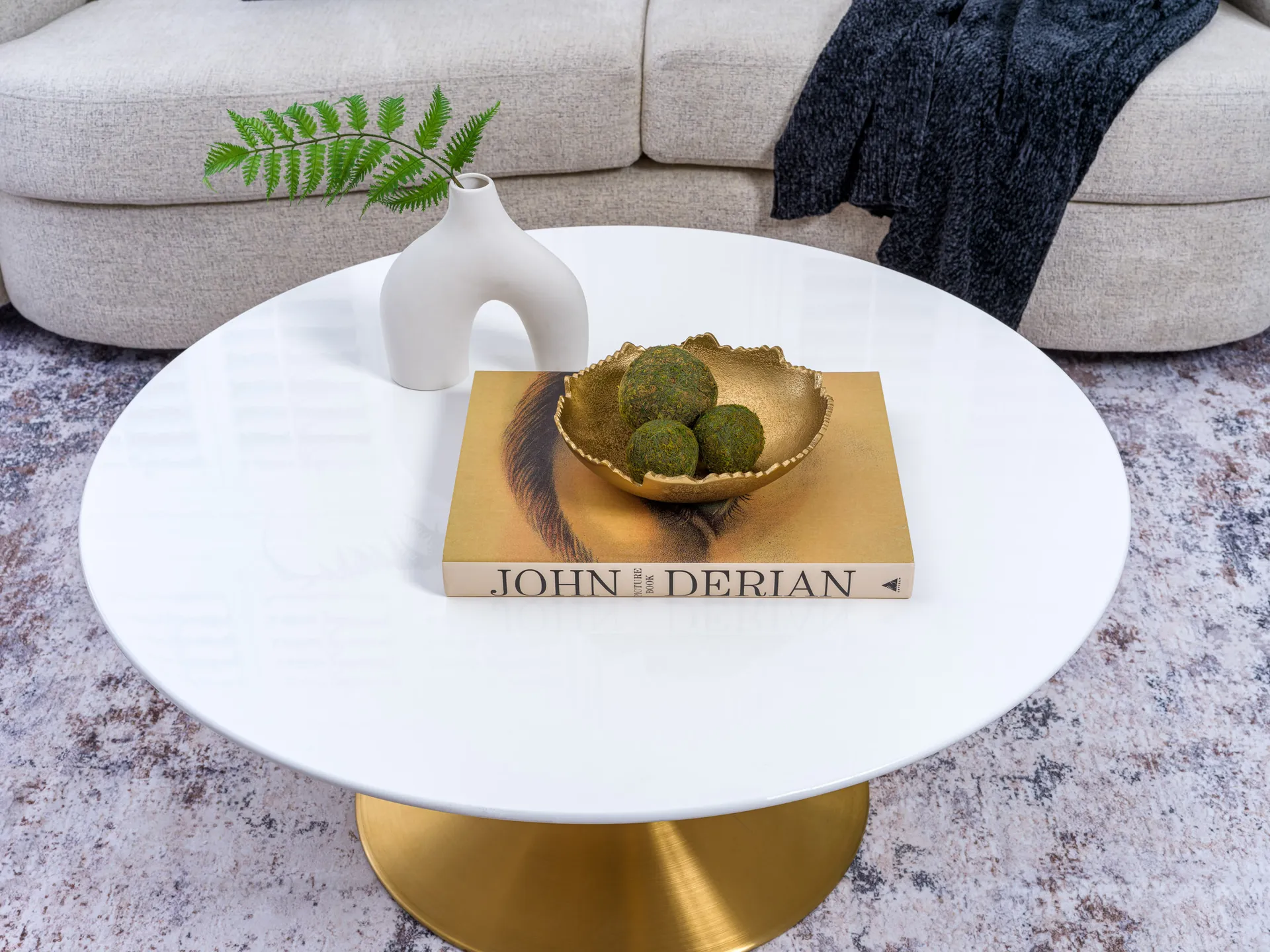A round white coffee table with a gold pedestal, featuring a gold bowl filled with moss spheres on a hardcover book, next to a sculptural vase holding a green fern.