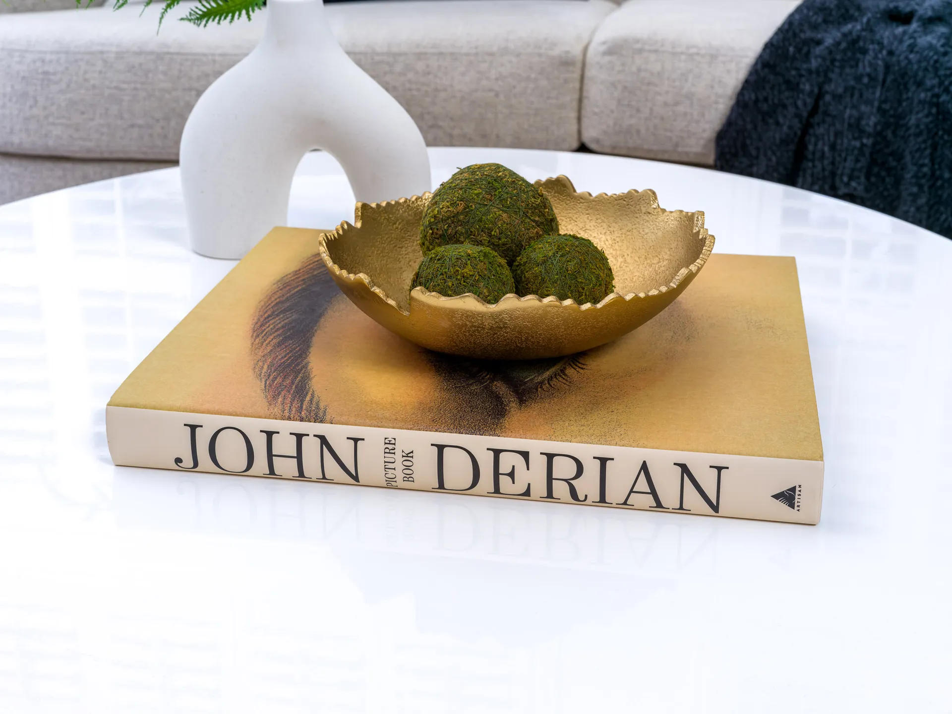 A scalloped gold bowl filled with moss spheres, resting on a hardcover book atop a glossy white table.