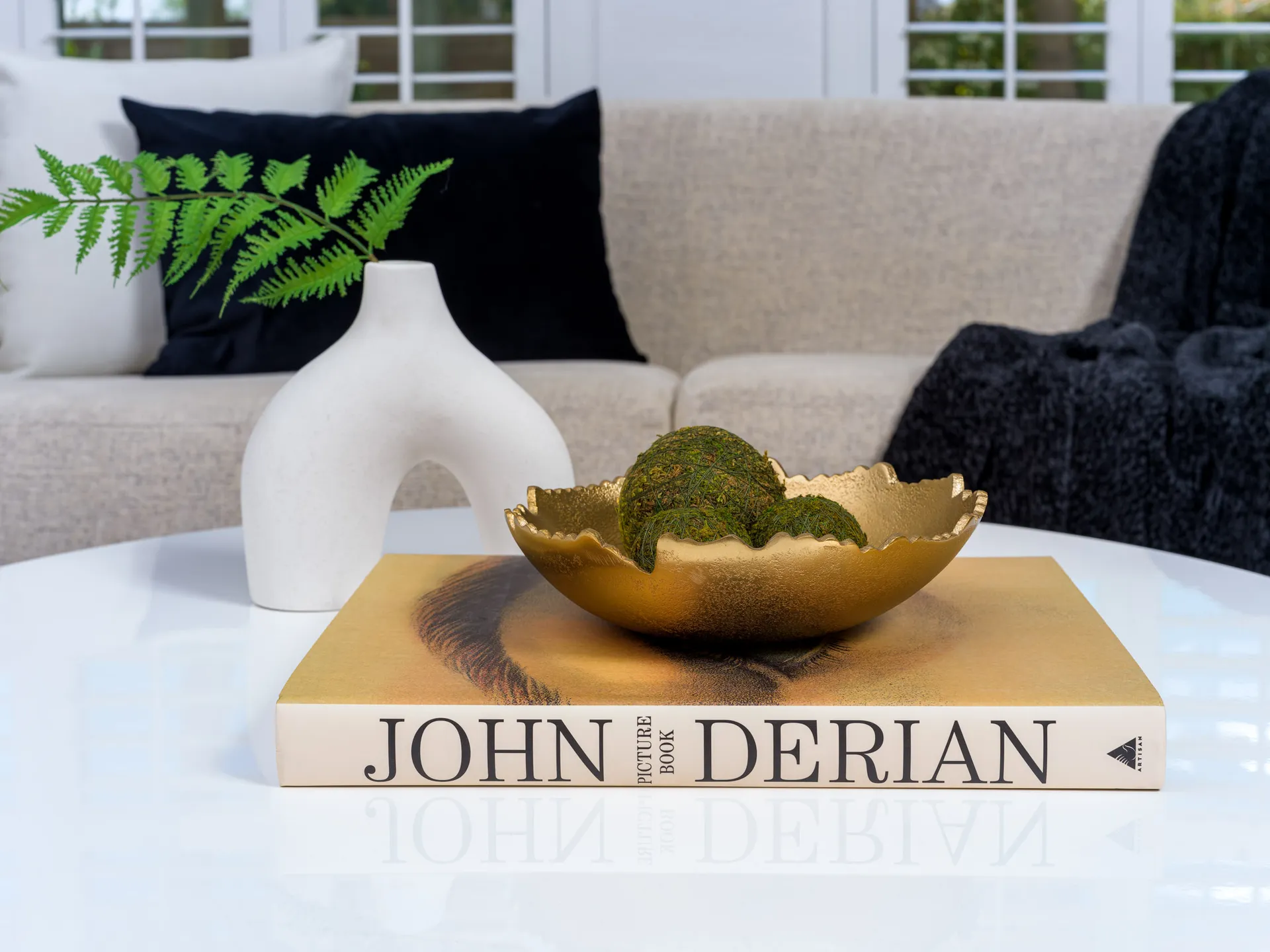 A white sculptural vase with a green fern and a gold bowl filled with moss spheres, set on a hardcover book atop a glossy coffee table.