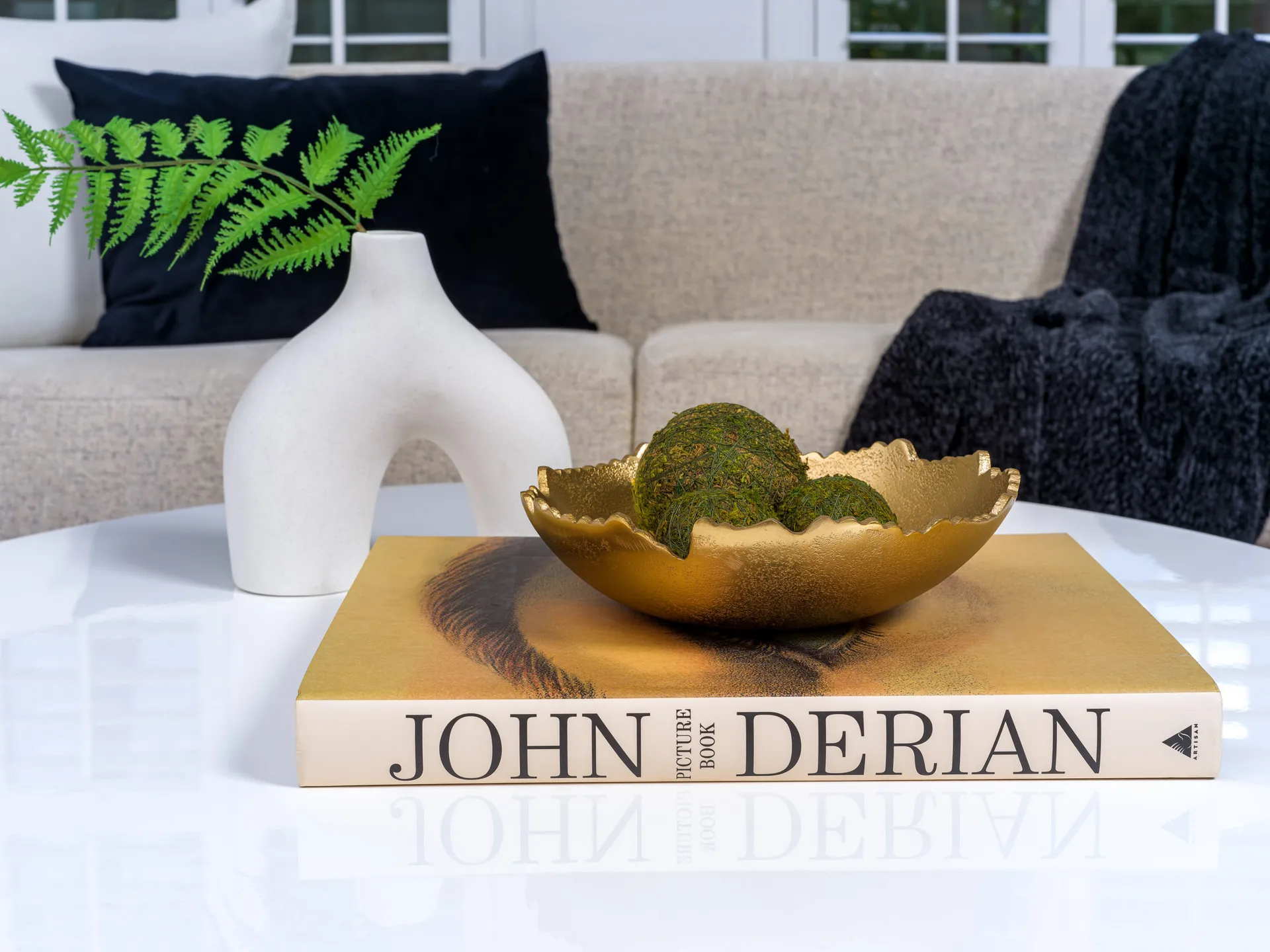 A gold bowl holding moss spheres on a hardcover book, alongside a white sculptural vase with a green fern on a glossy coffee table.