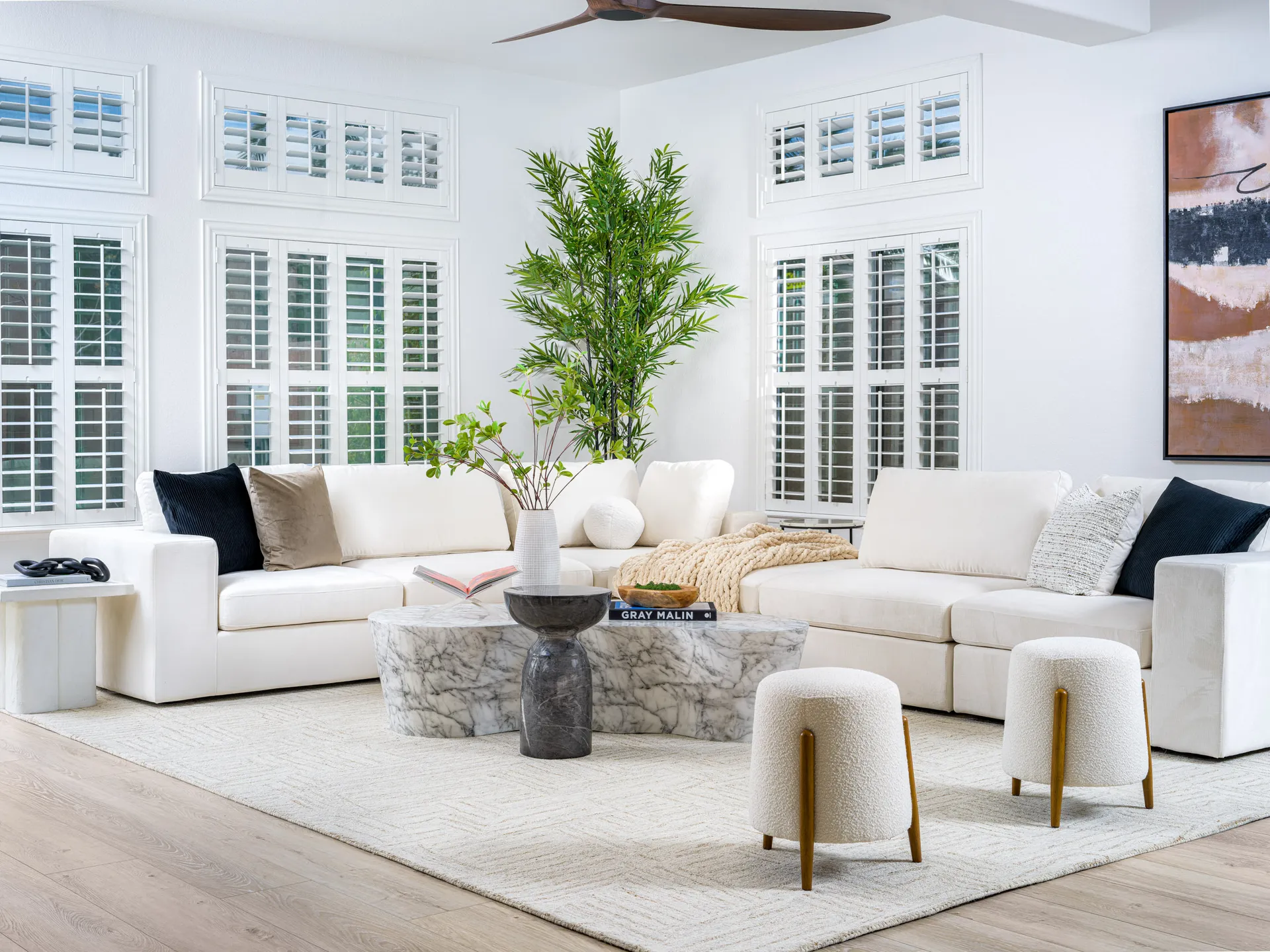 A spacious living room, styled by Contemporary Elegance Home Staging, featuring a white L-shaped sectional, a sculptural marble coffee table, two round stools with wooden legs, and tall shuttered windows.
