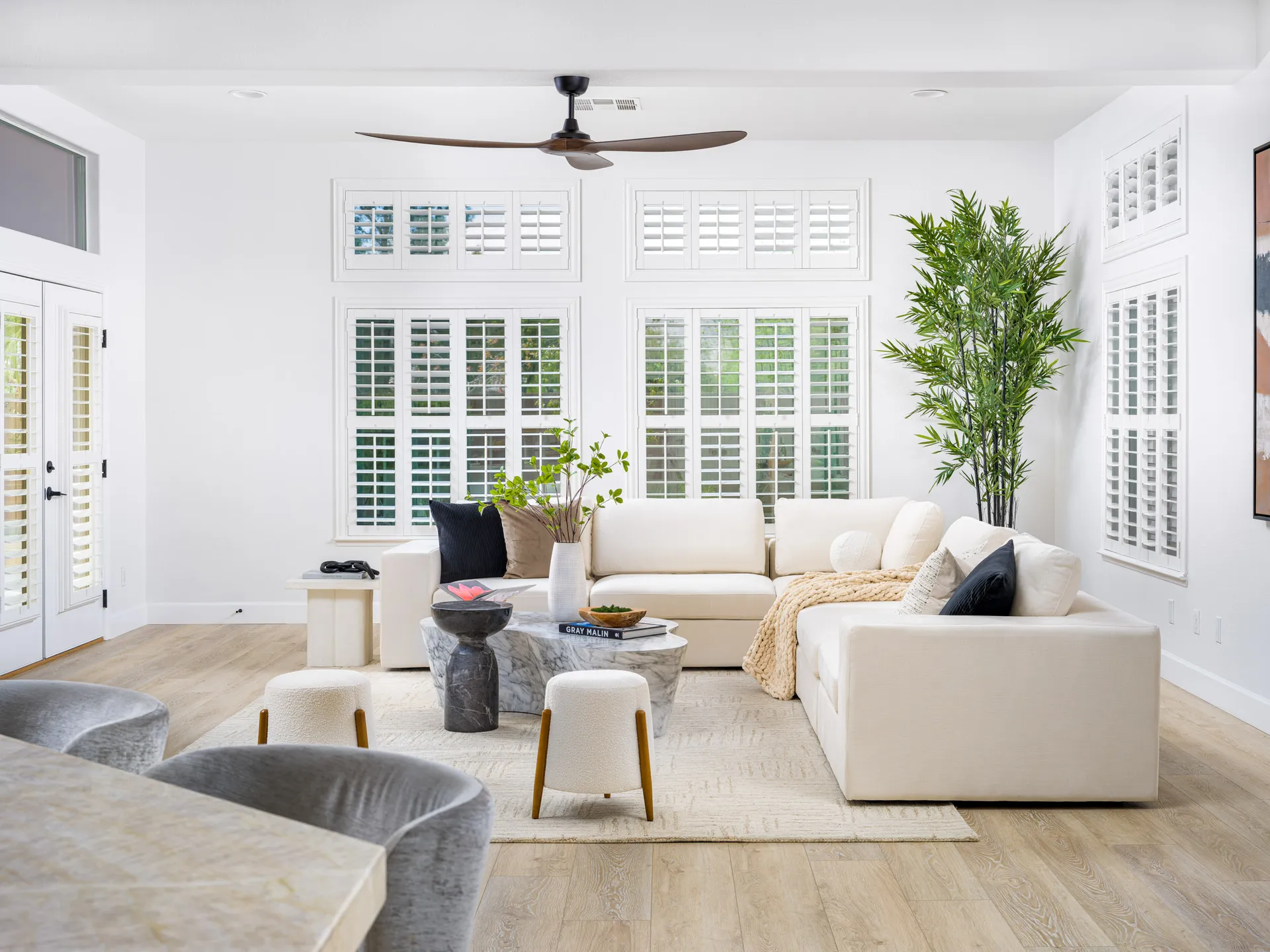 A bright living room featuring a white sectional sofa, a sculptural marble coffee table, tall shuttered windows, and a large potted plant.
