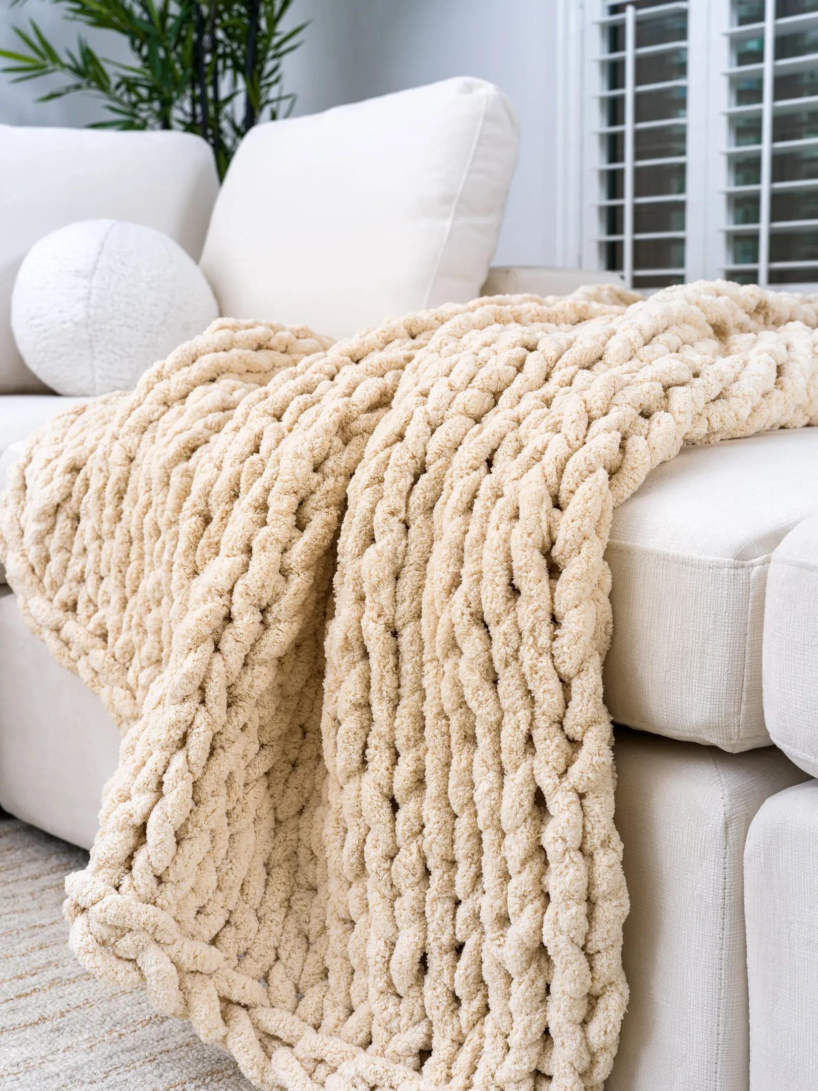 A thick, cream-colored chunky knit blanket draped over a light sofa.