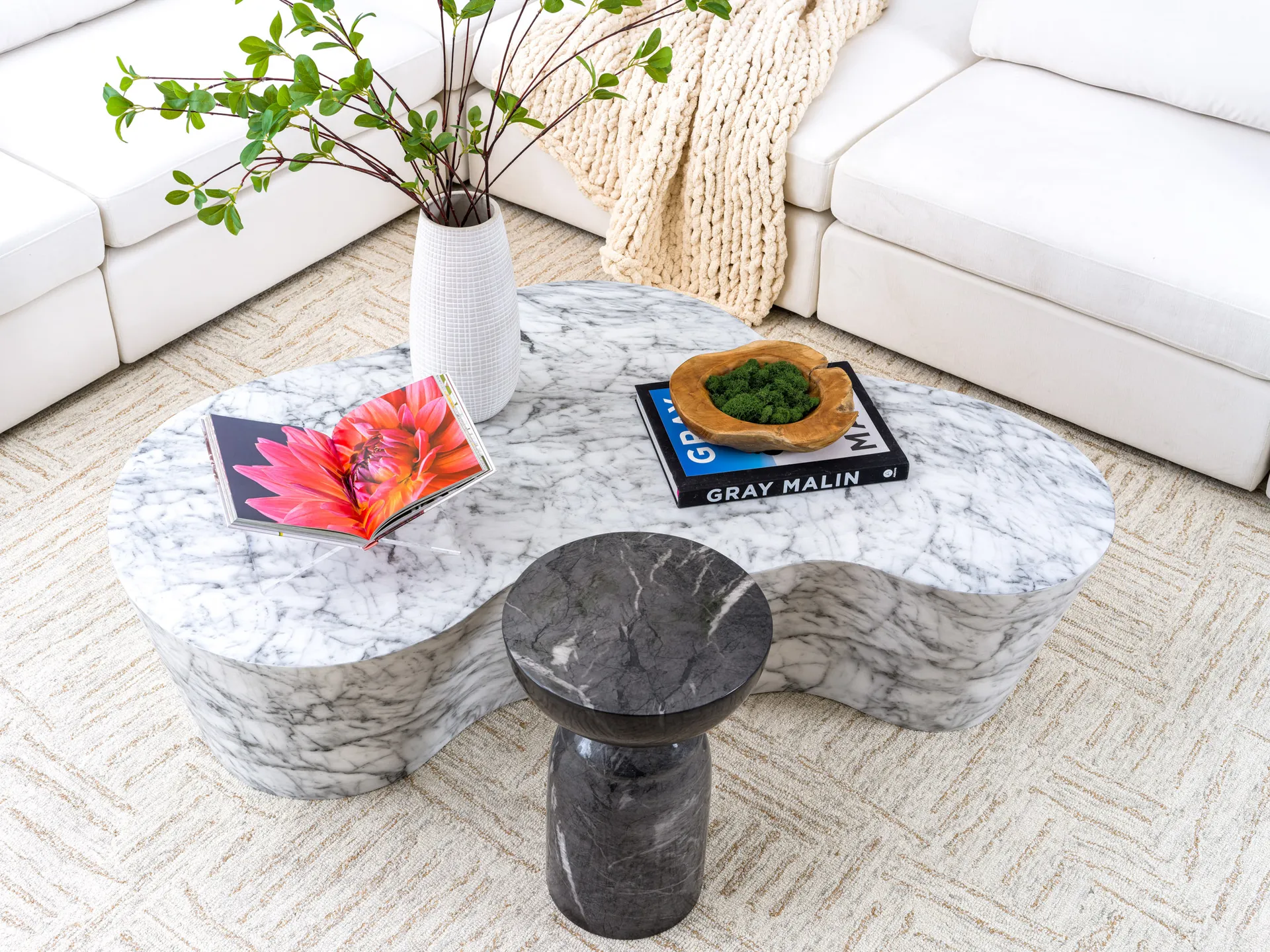 A curved marble coffee table displaying an open floral book, a wooden bowl of moss, and a white vase with greenery near a white sectional sofa.