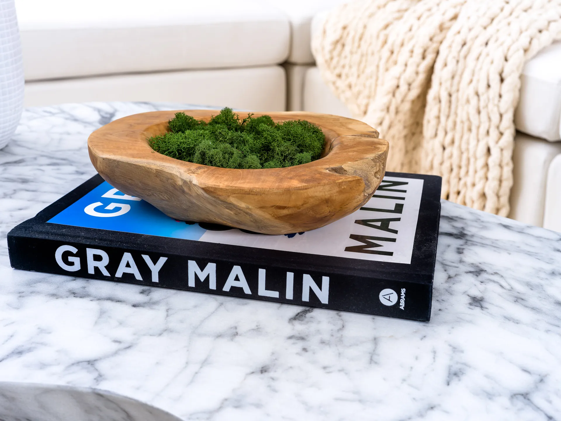 A wooden bowl with moss placed on a photography book on a marble coffee table.