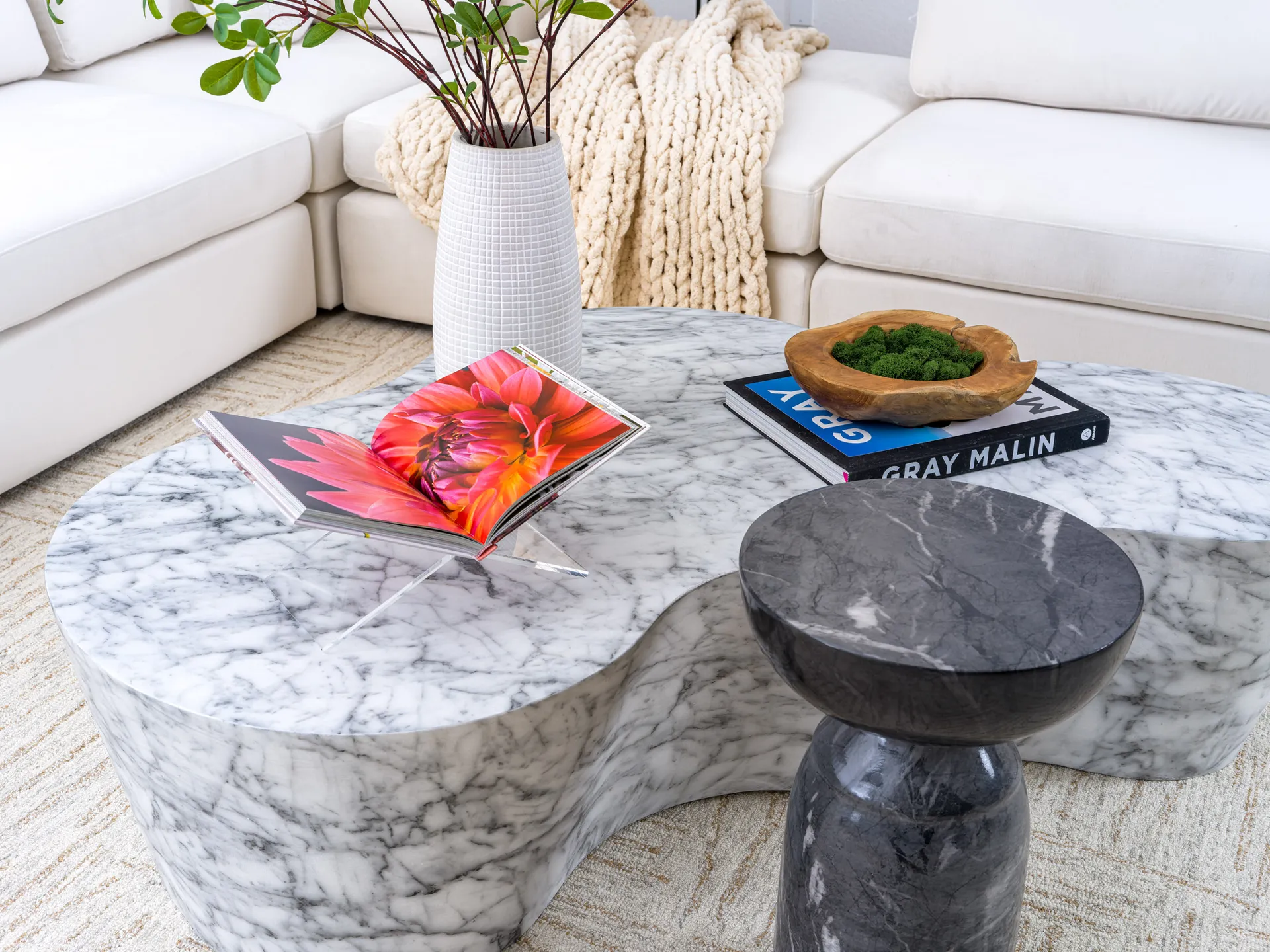 A curved marble coffee table displaying an open floral book, a wooden bowl of moss, and a white vase with greenery, next to a black side table.