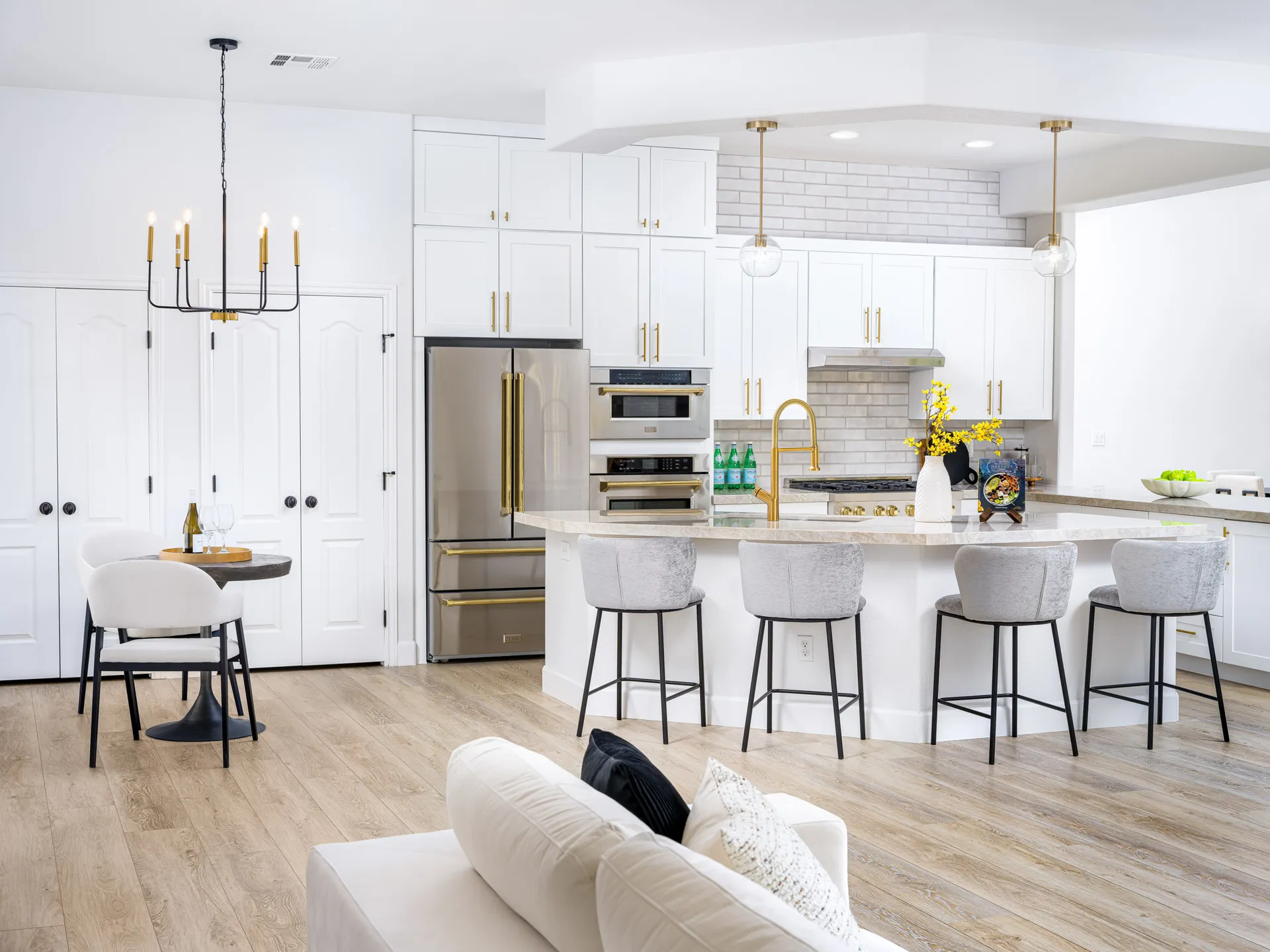 A bright white kitchen with gold hardware, an island with bar stools, and a small round table in the corner.
