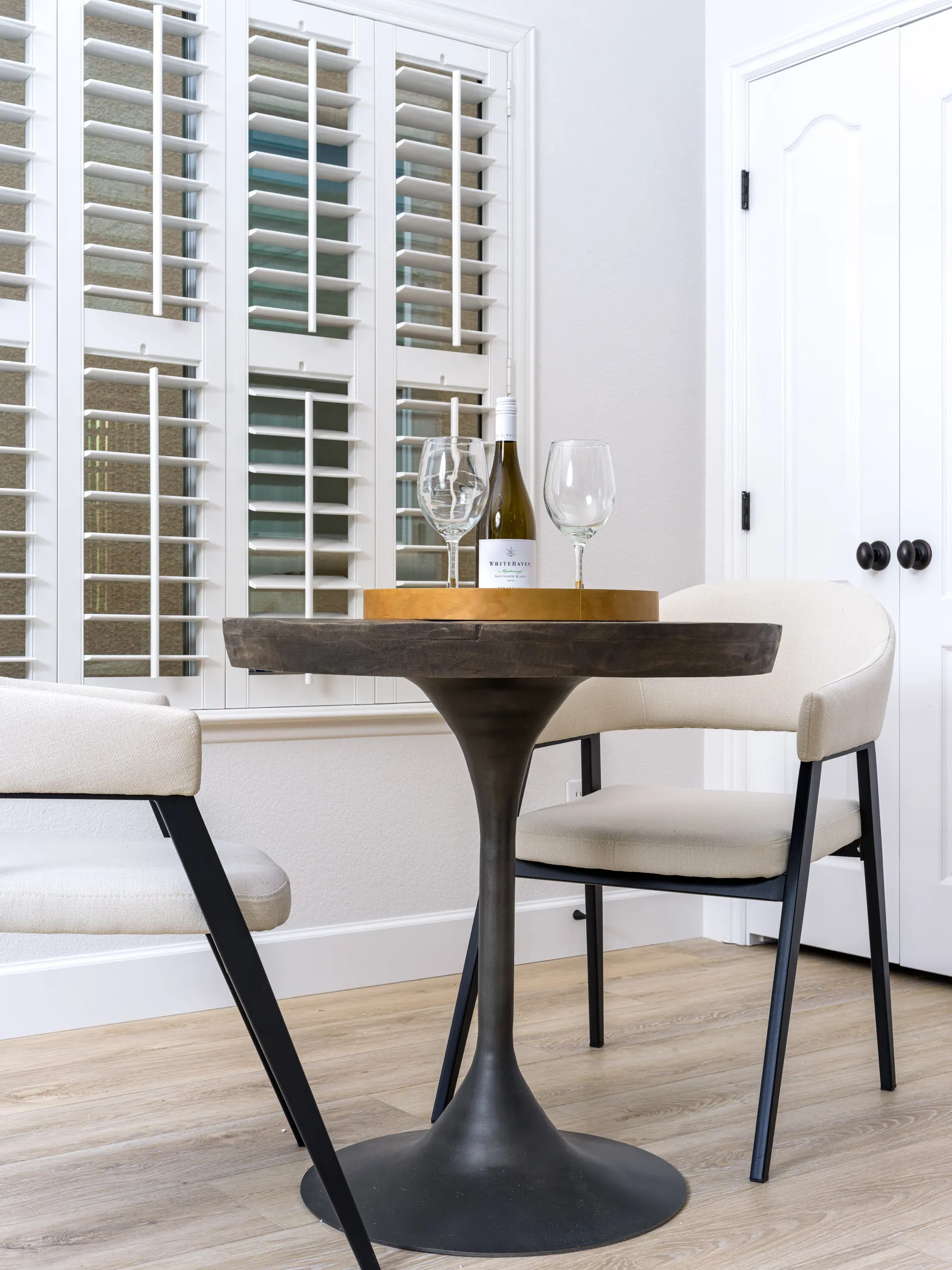 A round pedestal table with a mirrored tray holding a bottle of white wine and two glasses, flanked by modern chairs near white shutters.