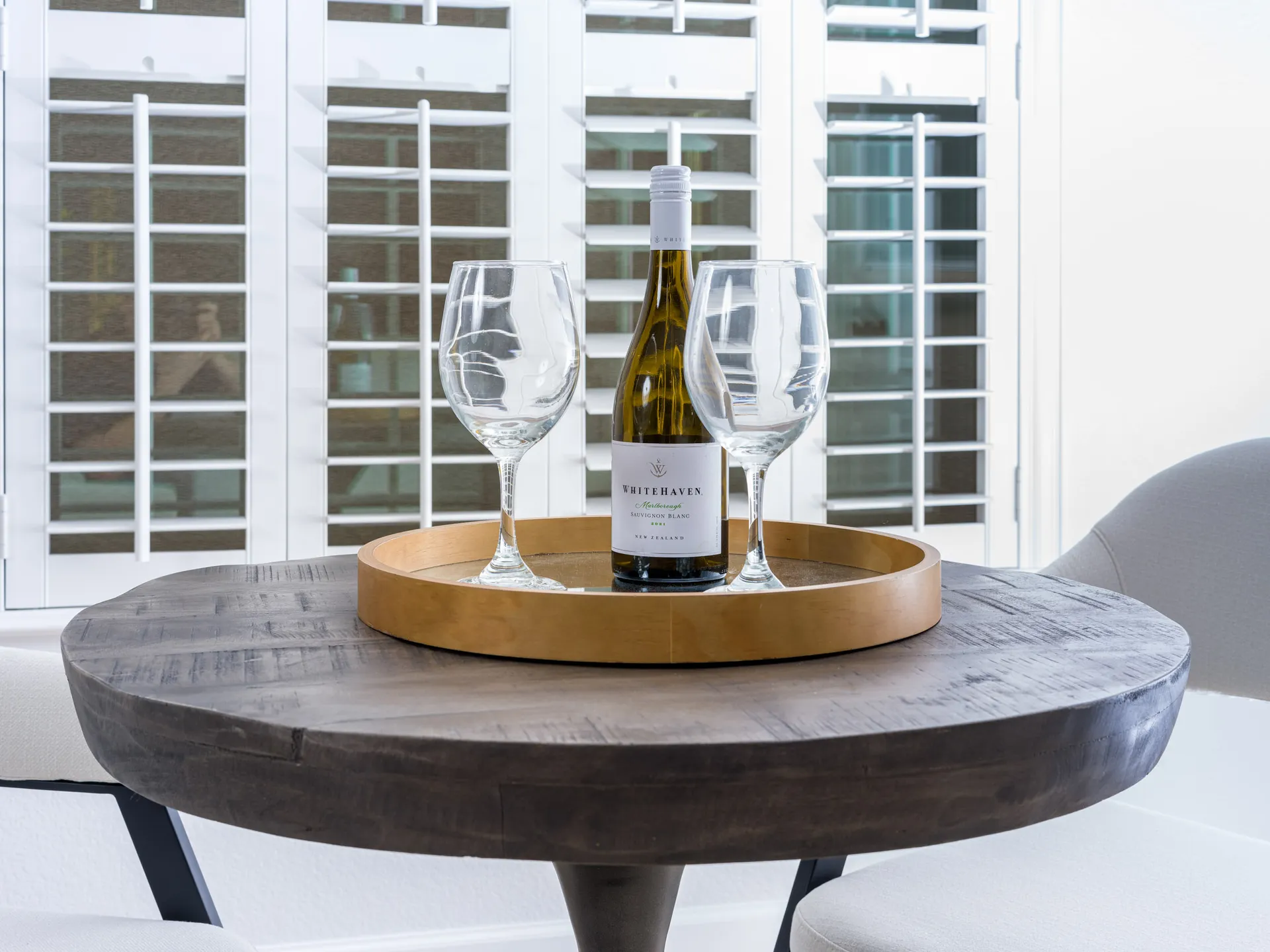 A round wooden table featuring a mirrored tray with a bottle of white wine and two glasses, in front of white shutters.