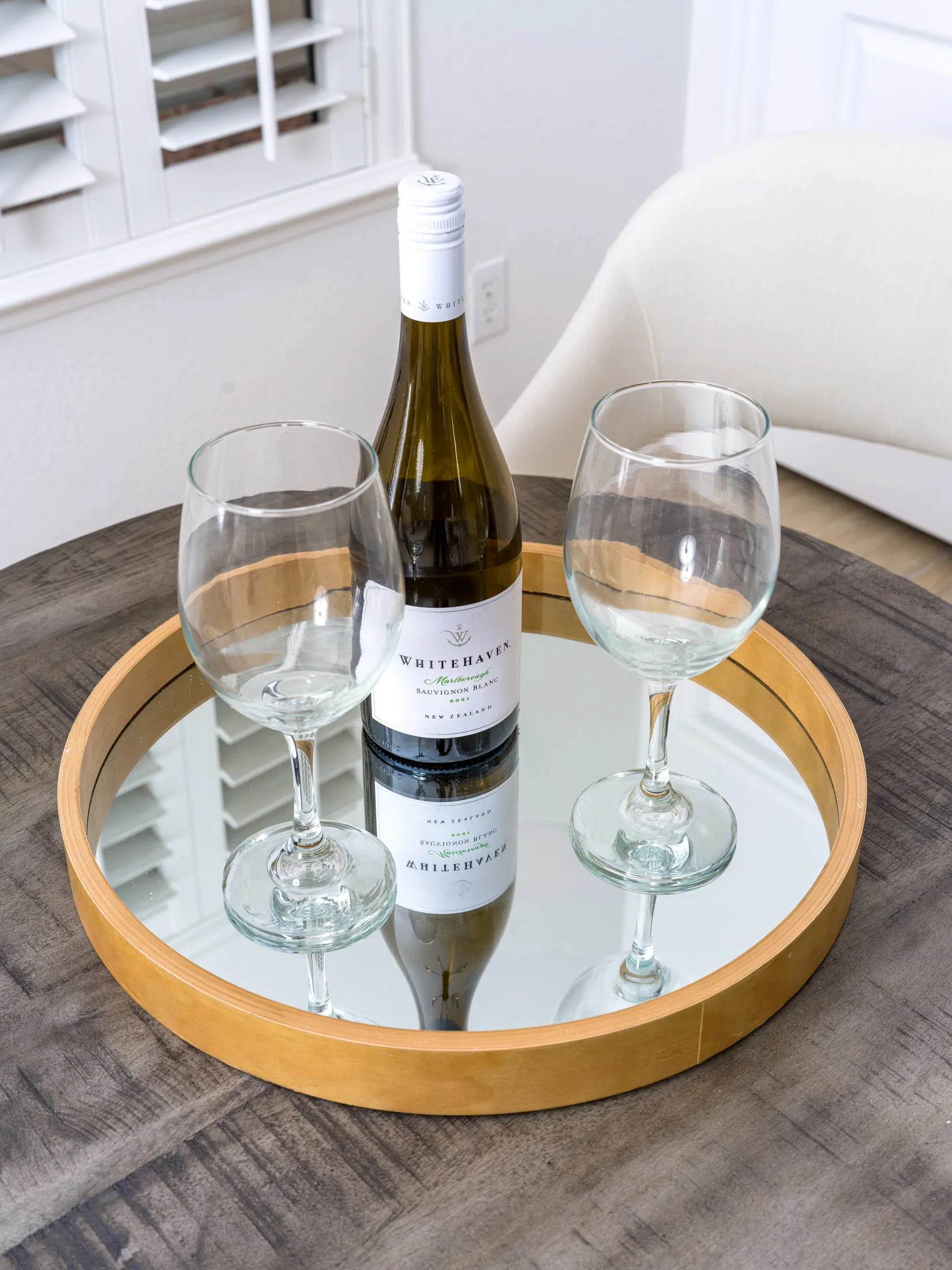 A round mirrored tray on a small wooden table, holding a bottle of white wine and two glasses.